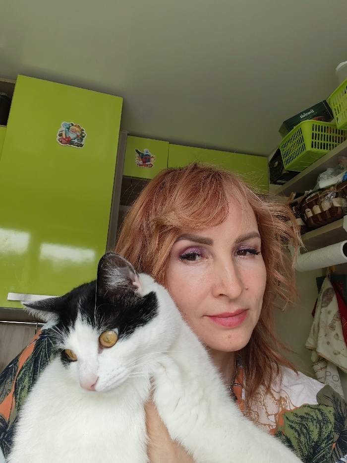 The image portrays a woman holding a cat in an interior setting, identified as being in the city of Chaikovsky, Russia. A woman with light reddish-brown hair, visible makeup including purple eyeshadow and pink lipstick, is positioned in the foreground, looking directly towards the viewer with a slight smile. She wears a patterned shirt featuring green and orange-brown leaf designs. She is holding a domestic cat, predominantly white with black markings on its head and ears, and possessing amber-yellow eyes. The cat is held against her left shoulder/chest, looking generally towards the left side of the frame. The background consists of light lime-green overhead cabinets with silver-colored handles. Two decorative magnets are affixed to the cabinet doors: one depicting a gnome-like figure with gifts on the left door, and another showing a bird on the right door. To the right of the frame, an open shelving unit is visible, containing light green plastic storage baskets, one of which holds a dark-colored box. Below the baskets, a roll of white paper towels is mounted on a holder, and various folded textiles are hanging. The upper wall and ceiling appear to be a light, neutral color.