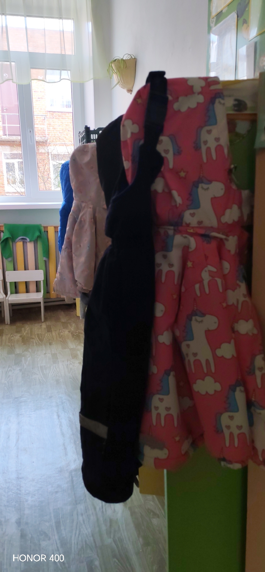 The image captures an indoor scene, likely a children's room, daycare, or kindergarten in Noviy, Russia, characterized by bright, natural daylight streaming through a large window on the left. The prominent subjects are several articles of children's clothing hanging on a rack or hooks. In the immediate foreground and right, a vibrant pink garment, possibly a dress or winter suit, features a pattern of white unicorns with blue manes and small white clouds. This garment is partially obscured by a dark blue or black puffy garment. Behind these, a light pink/peach puffy coat and a blue garment are also visible.
In the mid-ground on the left, two small, white wooden chairs sit beside a low, colorful wooden barrier, which has a green garment draped over it. The floor is light-colored wooden or laminate. Through the large window, brick buildings and bare tree branches are discernible, suggesting a late autumn or winter season in an urban or suburban environment. A sheer, light-yellow curtain hangs over the upper part of the window. On the white wall in the top right, a small wall-mounted planter holds a spider plant. The overall atmosphere is functional and child-oriented.
The only visible text is "HONOR 400" in the bottom left corner, likely a camera watermark. No specific activity is taking place; the scene depicts an organized space awaiting occupancy or use.
