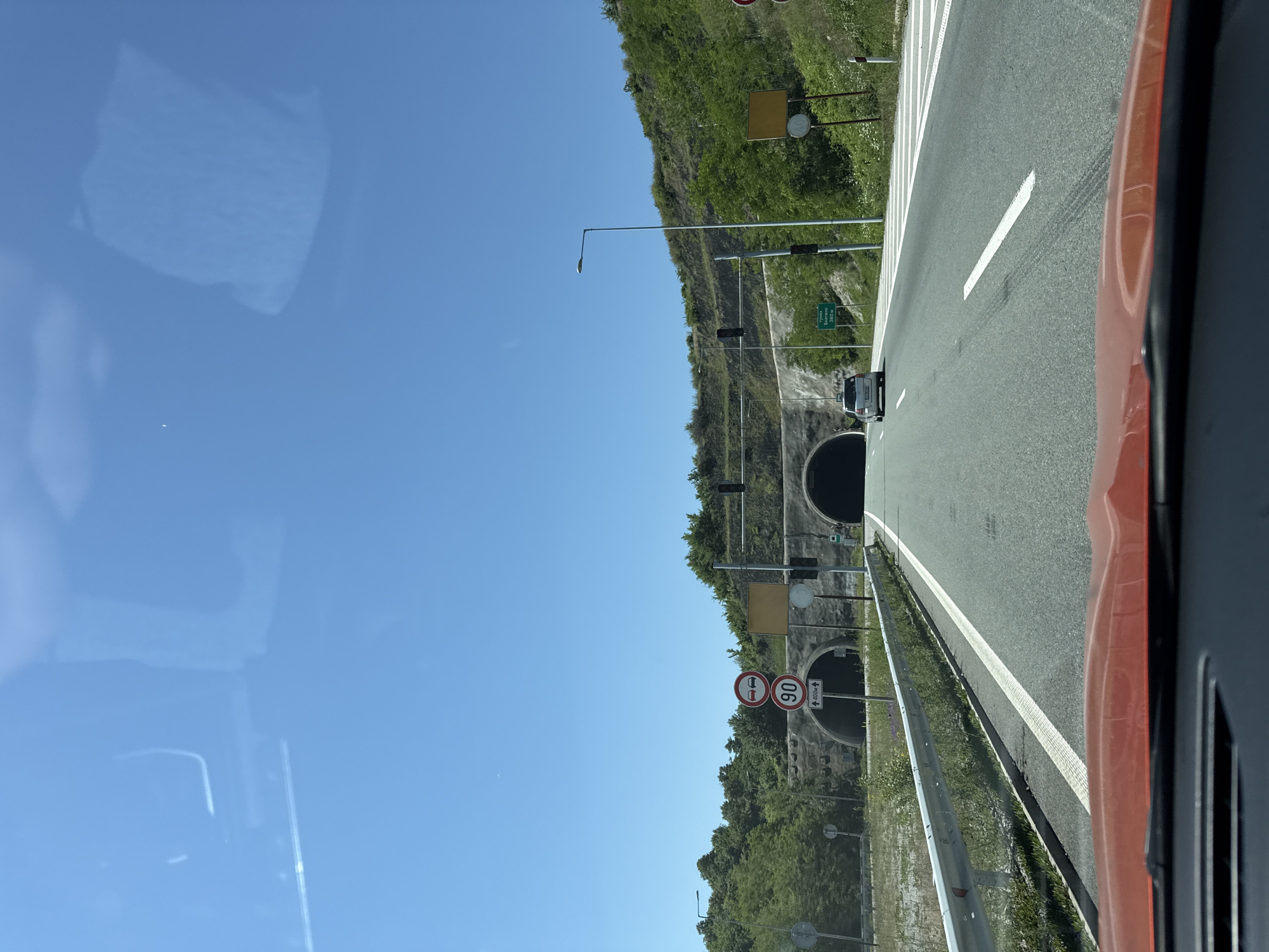 The image presents a forward-facing view from inside a vehicle on a multi-lane asphalt highway, approaching a tunnel system under a clear blue sky. The scene is illuminated by bright sunlight.
In the foreground, the top of an orange/red vehicle's dashboard and windshield is visible. The road ahead is well-maintained, featuring white dashed and solid lane markings. A silver hatchback car is visible in the distance, driving in the same direction towards the central tunnel entrance.
To the left of the road, a metal guardrail runs alongside a grassy embankment. Further left, two circular traffic signs are stacked vertically: the upper sign depicts two red cars, indicating a "no overtaking" zone, and the lower sign displays "90" in a red circle, denoting a 90 km/h speed limit. Below the speed limit sign, an upward arrow with "400m" indicates the duration or distance for which the limit applies or the tunnel length. Another smaller, circular white sign with a diagonal black line, likely signifying the end of a previous restriction, is partially visible.
Two tunnel entrances are visible, carved into a tree-covered hillside. The main tunnel is centrally located ahead, with another tunnel entrance appearing to its left. Both entrances are reinforced with concrete or stone structures. Above the tunnels, traffic signals with dark housings are mounted, likely controlling access or indicating tunnel conditions.
On the right side of the road, a green rectangular sign with white Cyrillic text is visible. It reads "ТУНЕЛА БЛАТИНО" (TUNELA BLATINO) and "380M", explicitly identifying the location as the Blatino Tunnel and stating its length as 380 meters. This places the scene at the Blatino Tunnel in Blatino, Bulgaria. Adjacent to the sign is a street lamp. Empty square yellow signs are present on both sides of the road near the tunnel entrances. The hillsides surrounding the road and tunnels are densely covered with green trees and shrubs.