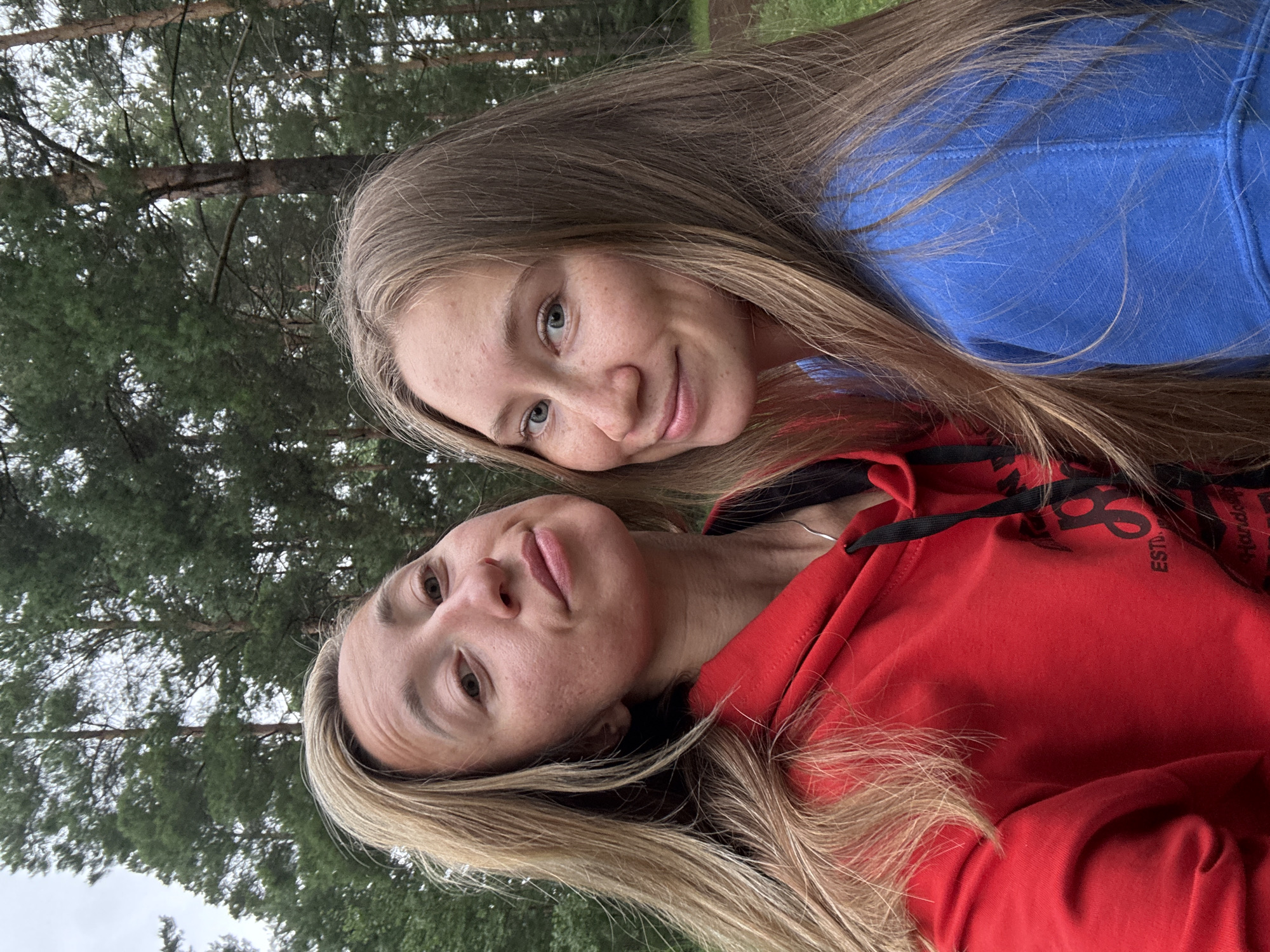The image is a selfie featuring two individuals, likely friends or family, posing for a photo outdoors. The scene is set amongst tall evergreen trees, suggesting a forest or park environment. The lighting indicates it is daytime, possibly overcast or late afternoon, as there are no harsh shadows.
The person in the foreground, wearing a red hoodie, is looking directly at the camera with a neutral expression. Their blonde hair is visible framing their face. Behind them, another person, wearing a blue hoodie, is partially visible. This individual also has blonde hair and is smiling at the camera, with their eyes conveying a sense of happiness.
There is some visible text on the red hoodie, appearing to be a logo with letters, including "ESTD." and possibly "Florida" although the latter is partially obscured and difficult to read definitively. The overall mood of the image is casual and friendly, capturing a moment of shared activity in a natural setting. Given the location context of Прикамский, Russia, the trees likely consist of native species common to that region.