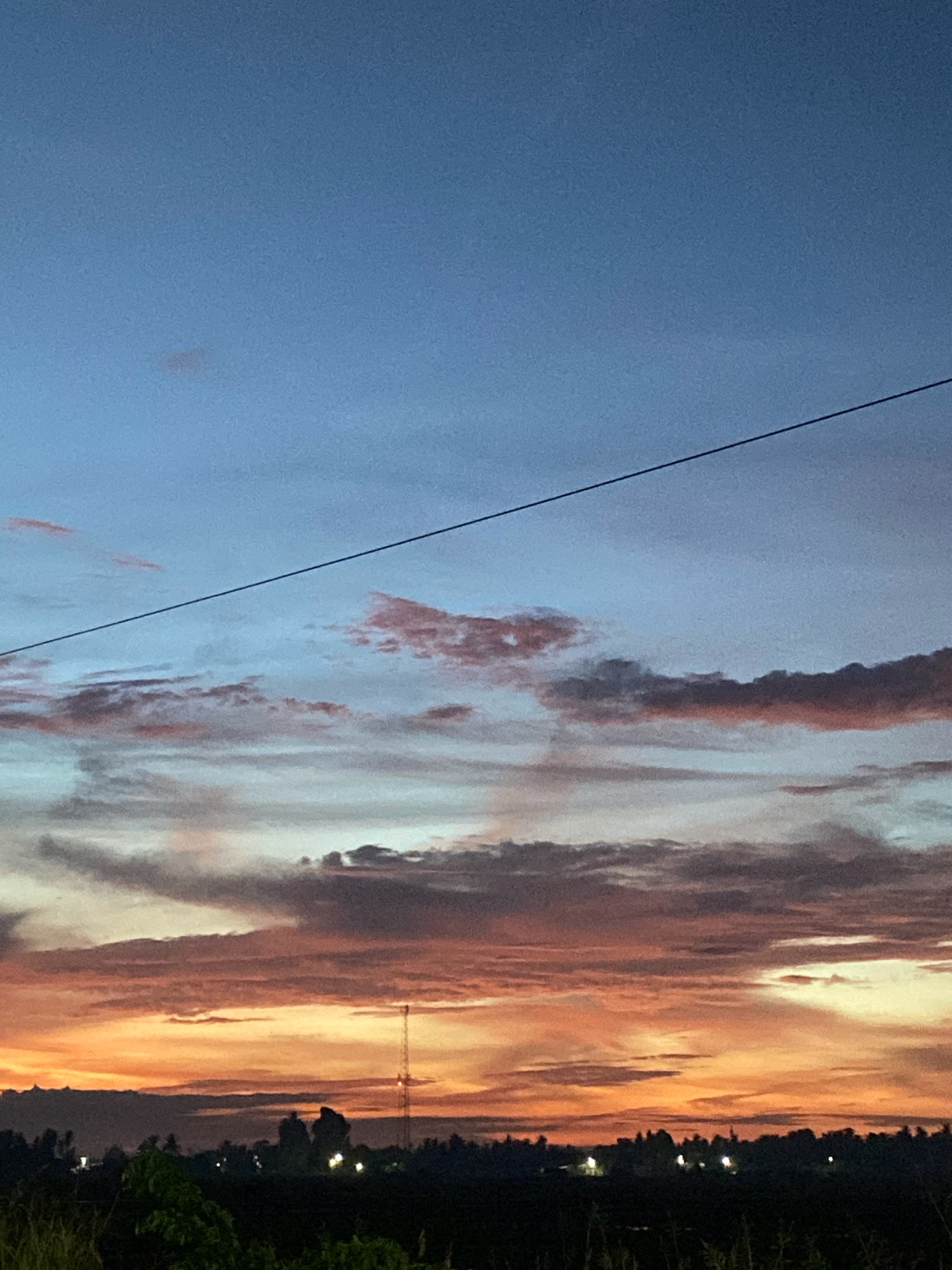 This is a landscape photograph of a sunset over a rural area, likely in Cotgapu, Indonesia. The sky is a gradient of deep blue at the top, transitioning to lighter blues and then fiery oranges and pinks near the horizon. Wispy clouds are scattered throughout the sky, catching the colors of the setting sun. A thin, dark power line stretches diagonally across the upper half of the image.
In the lower portion of the image, a silhouetted tree line marks the horizon. A tall radio tower with lights is visible on the right side of the tree line. Several other small lights are scattered along the horizon, suggesting a small settlement or scattered homes. The foreground is dark and indistinct, appearing to be fields or vegetation. There are no people or animals clearly visible in the image. The overall impression is one of a peaceful, natural scene at dusk.
