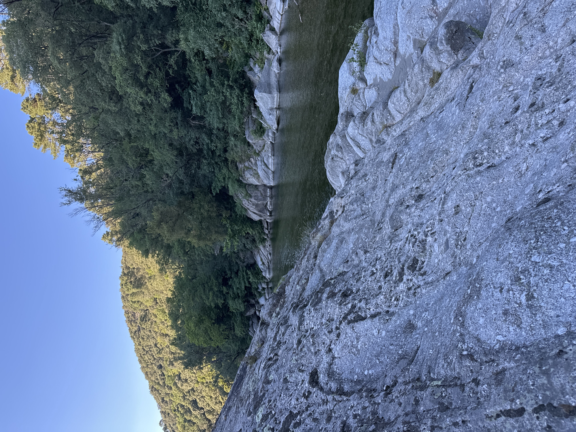 The image depicts a natural scene featuring a riverbank and a wooded hillside. In the foreground, large, textured grey rocks dominate the scene, sloping downwards towards the water. The river itself is calm and dark, with its surface reflecting the surrounding foliage. Along the opposite bank, a dense forest covers a steep hillside. The trees are predominantly green, with some appearing taller and more mature, possibly pines, interspersed with smaller deciduous trees. The sky above is clear and blue, indicating fair weather. The light suggests it is either early morning or late afternoon, with the sun casting shadows and highlighting the textures of the rocks and the canopy of the trees. The location is identified as being in the vicinity of Anduze, France.