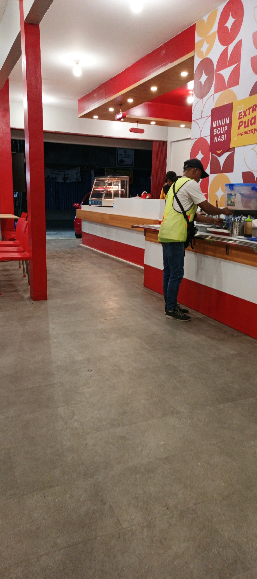 The image shows the interior of a fast-food restaurant, likely a franchise, in Sidoarum, Indonesia. Red and white are the dominant colors in the decor, with red pillars and a red and white striped counter. The walls feature a repeating geometric pattern in red and yellow, along with signage for "MINUM SOUP NASI" and "EXTRA PUAS... sepuasnya".
In the foreground, a man wearing a yellow safety vest over a white shirt and blue jeans stands behind the counter, appearing to be serving customers or preparing food. He is wearing a dark baseball cap. To his left, another person, possibly a customer or employee, is partially visible.
Further back, a glass display case is visible on the counter, likely for food items. A red car is parked outside the establishment, visible through an open doorway or large window in the background. The floor is concrete. The lighting suggests it is evening or indoors with artificial illumination.