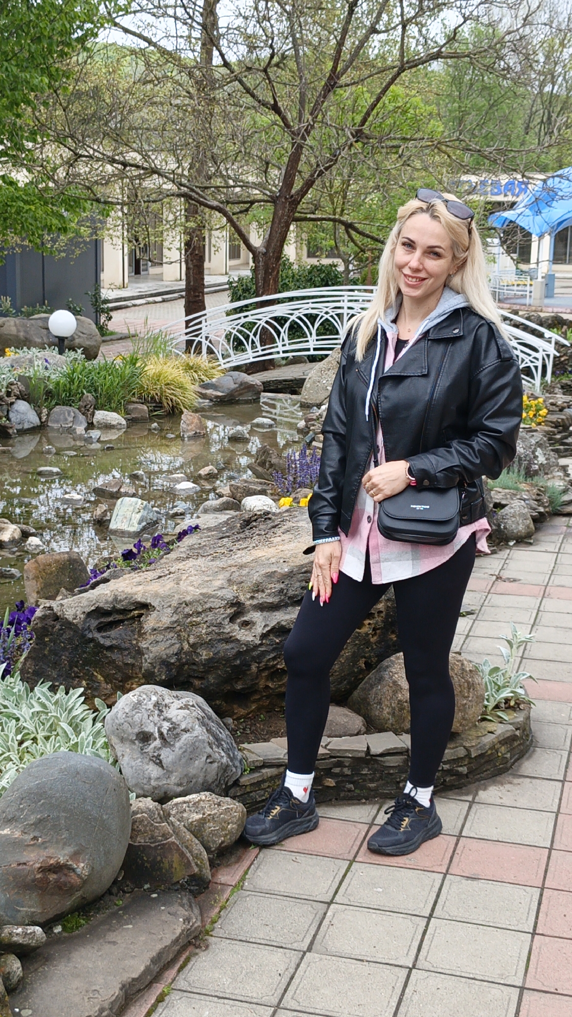 A blonde woman stands on a tiled walkway in Goryachiy Klyuch, Russia, next to a pond with rocks, plants, and a small white bridge. She wears a black leather jacket, a plaid shirt, black leggings, and black sneakers with white socks. A black purse hangs from her shoulder. Sunglasses rest on her head. The background includes trees, buildings, and a partially visible blue awning.