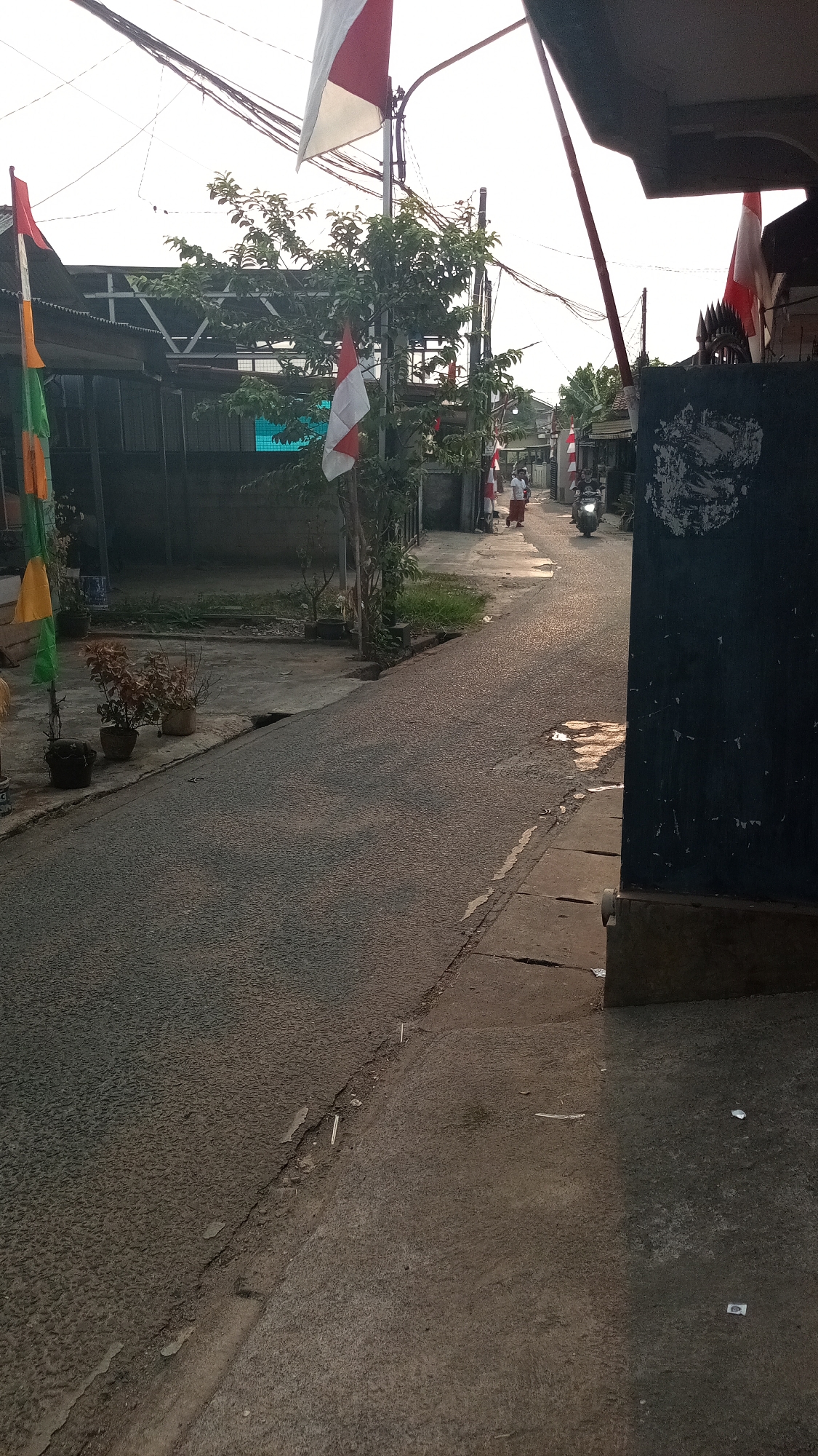The image depicts a narrow, paved street in a residential area in Depok, Indonesia, during daylight, likely late afternoon due to the lighting. The street is bordered by modest buildings with dark facades on the right and a lower concrete area with plants and houses on the left. Several Indonesian flags, red and white, are displayed on poles along the street, suggesting a national holiday or celebration. Two individuals are visible walking down the street in the distance, and a motorcycle is being ridden towards the viewer. The road surface is textured and shows signs of wear and some puddles reflecting the light. Overhead, a tangle of electrical wires crisscrosses the sky.