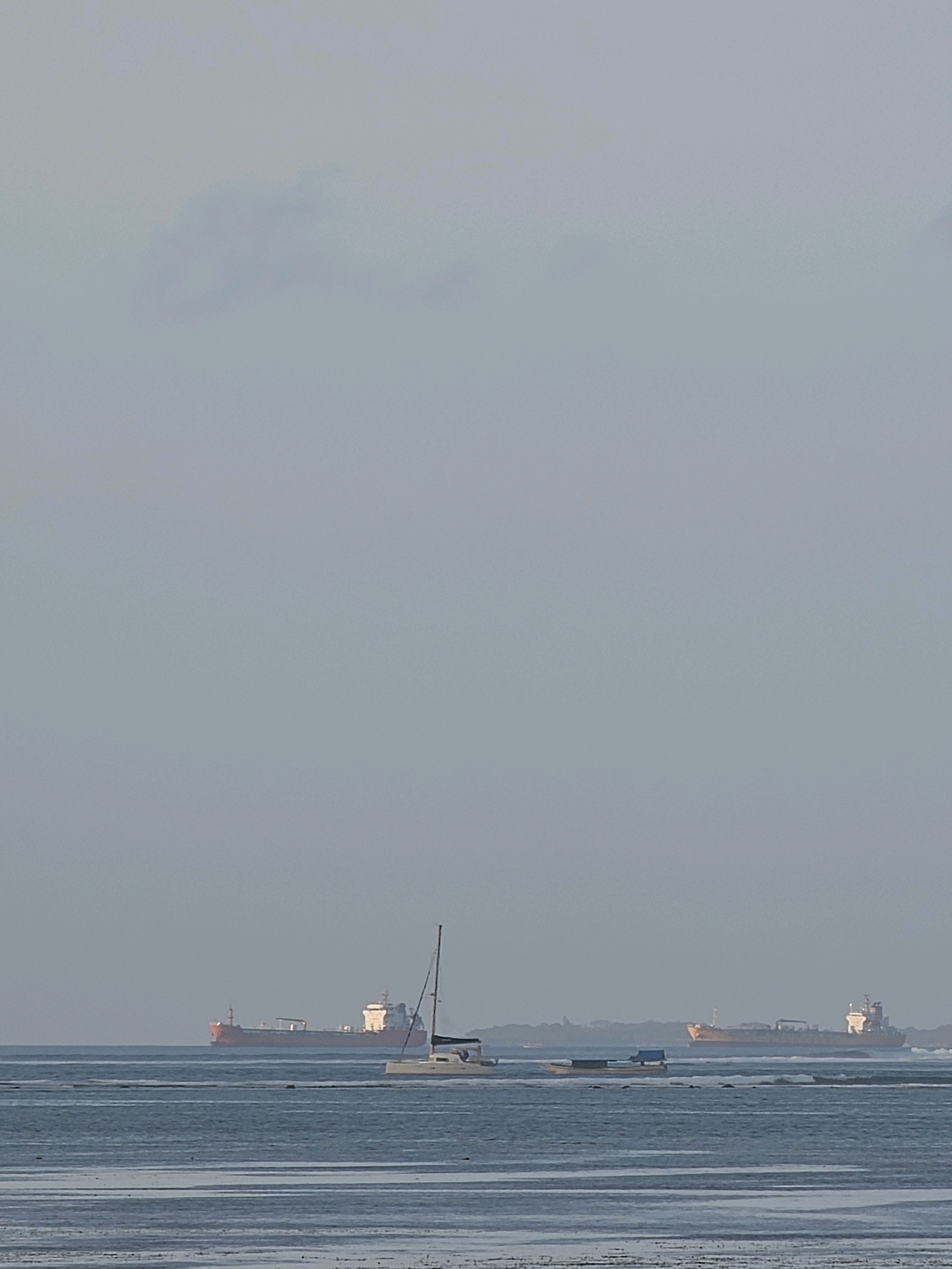 The image depicts a body of water with several vessels under a hazy, overcast sky. In the distance, two large cargo ships are visible. One is a reddish-orange vessel on the left, and the other is a lighter orange vessel on the right. Closer to the foreground, a white sailboat with a single mast is present. Between the sailboat and the right cargo ship, two smaller, elongated vessels with blue coverings are also in the water. The water itself appears calm with some ripples, and the foreground shows a wet, sandy or muddy shoreline. The scene suggests a port or coastal area, potentially near Denpasar, Indonesia, given the presence of cargo ships and the tropical haze. There are no people clearly visible in the image.