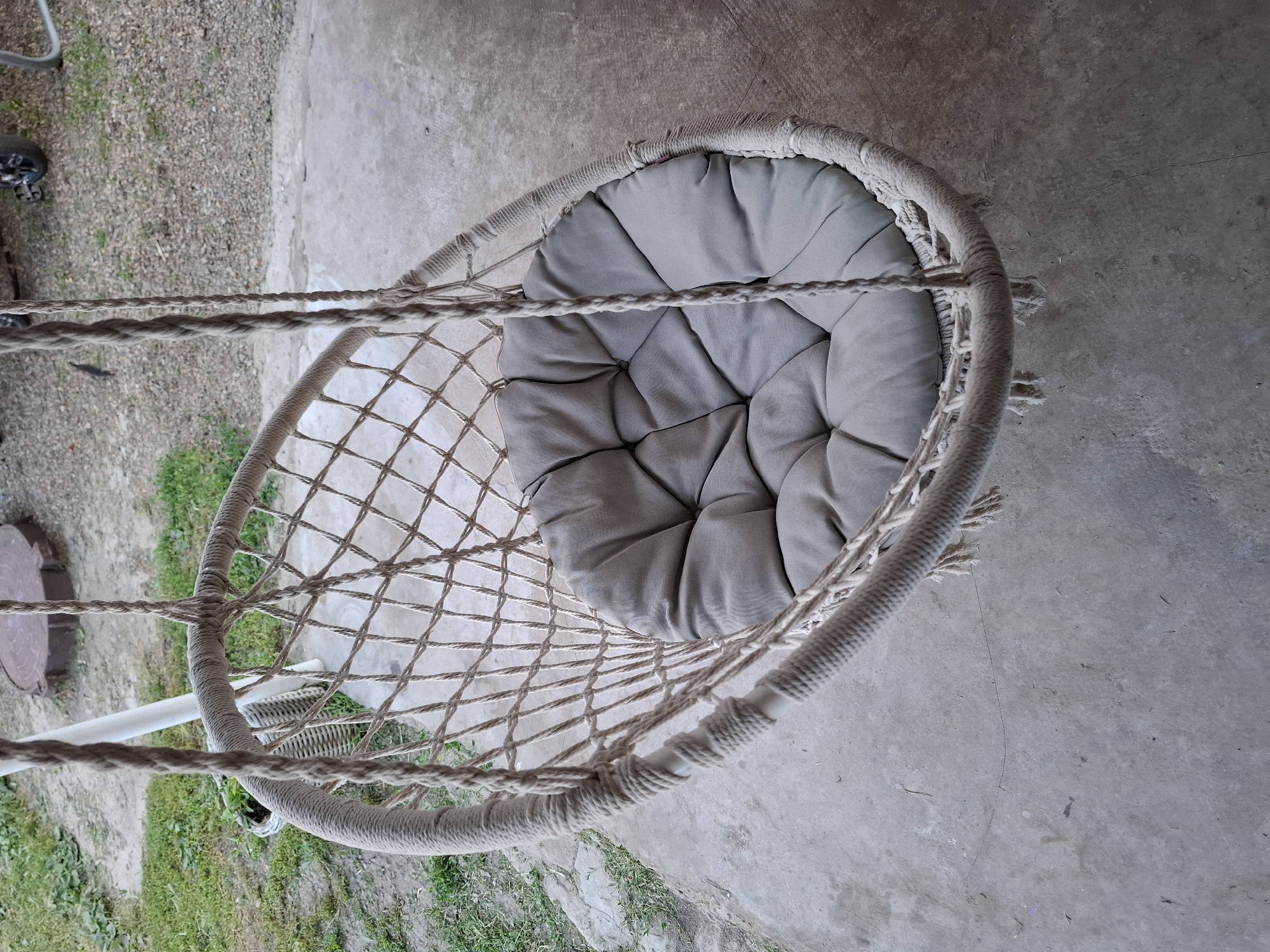 A hanging chair with a grey cushion is suspended by ropes in front of a concrete patio. The chair is made of woven rope with a diamond pattern and fringe details. In the background, there is green grass, a white pole, and a large circular object that appears to be a manhole cover. The ground is cracked concrete with some gravel and debris. The scene is outdoors and appears to be in a residential or outdoor recreational area.
