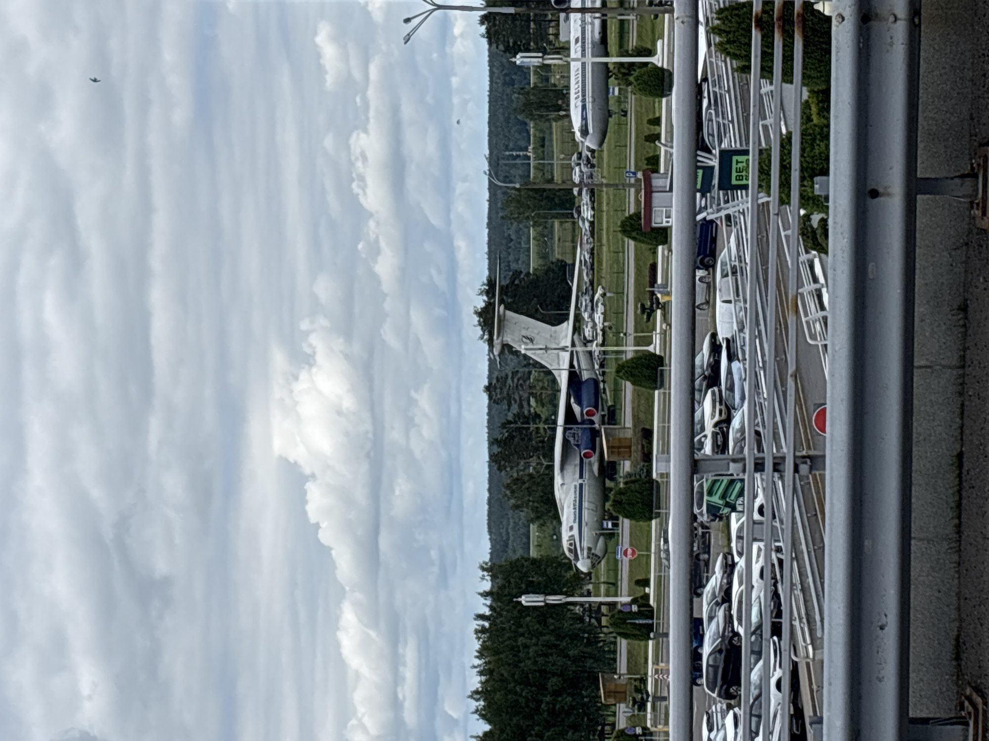 The image depicts an outdoor scene with an overcast sky, featuring two large airplanes parked on a tarmac. In the foreground, there is a parking lot filled with various vehicles. A metallic railing runs horizontally across the bottom of the frame. Behind the parking lot, a line of dense green trees forms a backdrop, with more distant, undulating green terrain beneath the sky. The environment appears to be an airport or an aviation museum, potentially in Minsk, Belarus, given the presence of a Belarusian flag on one of the aircraft and a sign with "BELARUS" visible.