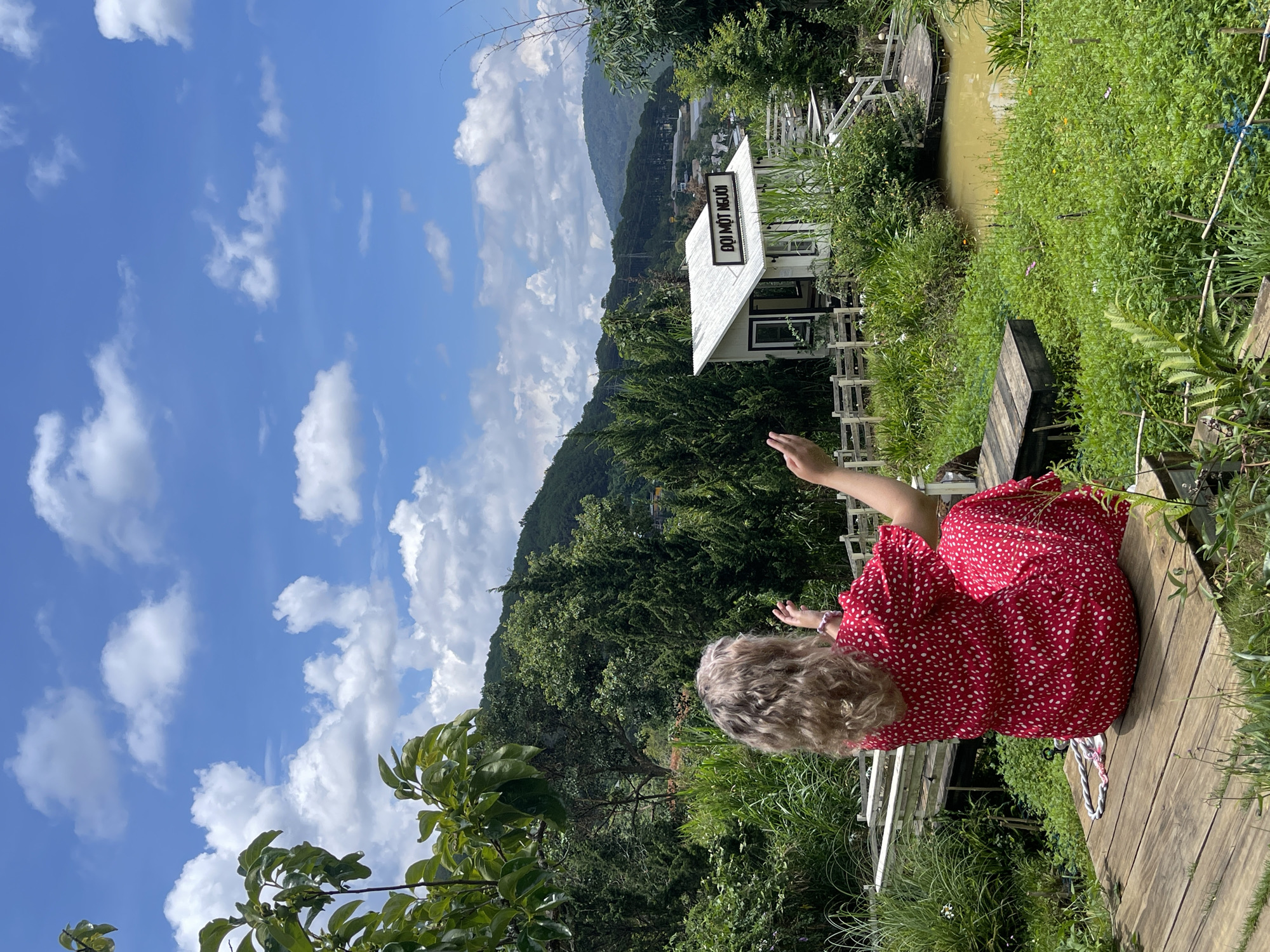 A woman in a red and white patterned dress sits on a wooden path, extending her right arm upward. The path is surrounded by green vegetation and leads to a small white building with a black sign on top, indicating "DOI MOT NGUOI". Behind the building is a body of water, likely a canal or small river. Further in the background are green mountains under a blue sky dotted with white clouds. The scene is likely set in Da Lat, Vietnam.