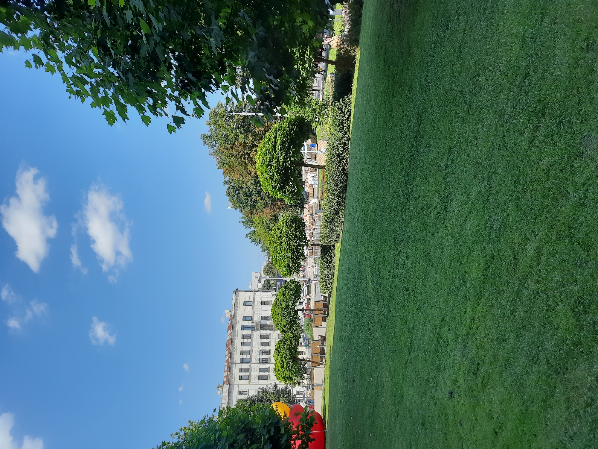 A bright, sunny day in Lviv, Ukraine. A lush green lawn dominates the foreground, stretching into the distance. Beyond the lawn, a line of neatly trimmed trees and bushes separates the park from a city street. In the background, a multi-story building with a light-colored facade stands, its windows reflecting the sunlight. A few scattered clouds drift across the clear blue sky. Red and yellow spherical objects are visible in the lower left corner, possibly part of an art installation or playground equipment. The scene conveys a sense of peaceful urban greenery, perfect for a leisurely afternoon.