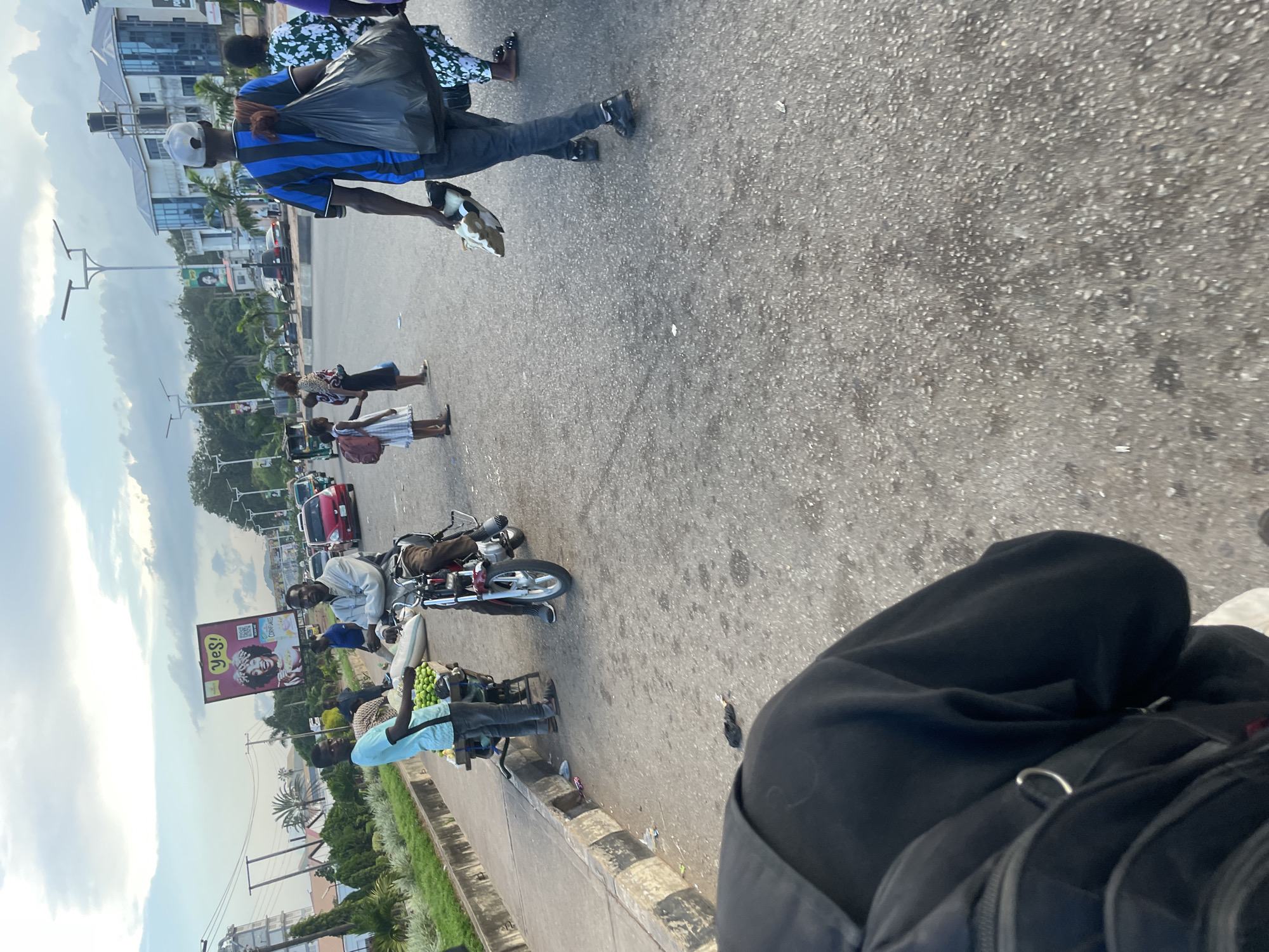 The image depicts a street scene in Barakin Lerawi, Nigeria, likely during daytime given the lighting. The foreground is dominated by the back of a person wearing a black hoodie and a backpack. The middle ground features a street with several individuals and vehicles. A man is seated on a red motorcycle, appearing to be speaking with a younger man standing next to a cart laden with green produce, likely limes. Other people are walking along the street, including a man in a blue and black striped shirt carrying a large black plastic bag and another person holding a pair of shoes. In the background, there are buildings, trees, a billboard with the word "YES!" on it, and streetlights. Traffic, including cars and what appears to be a tricycle-like vehicle, is visible on the road. The overall environment suggests a busy urban setting.