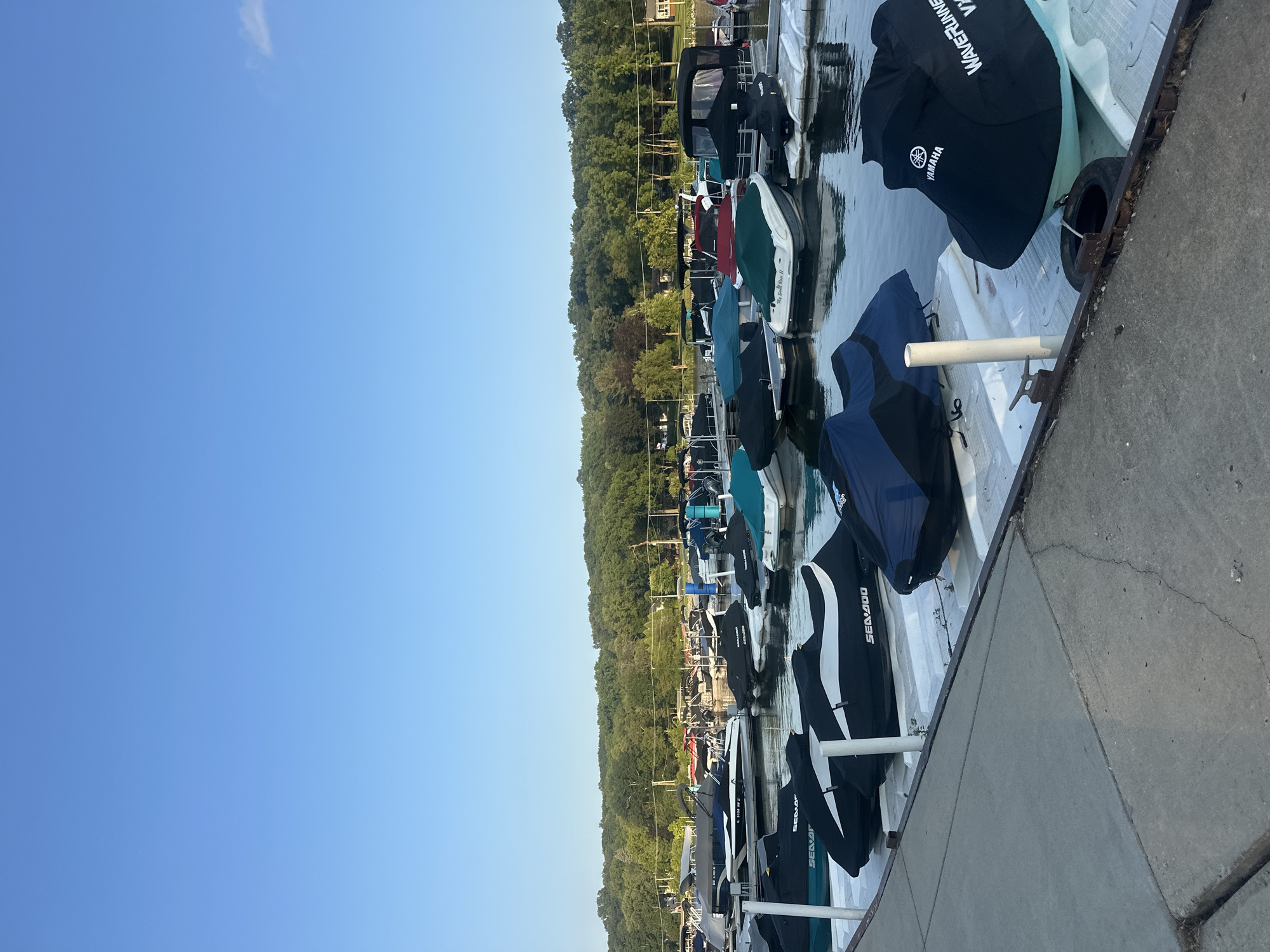 The image shows a marina on a body of water, likely a lake, in Algonquin, Illinois, United States of America. The sky is clear and blue with a hint of a distant cloud. The water is calm and reflects the sky and surrounding trees. In the foreground, a concrete walkway runs along the edge of the marina. Moored in the water are numerous personal watercraft (jet skis) and boats, most of which are covered with protective tarps. Some of the jet skis have visible branding, such as "SEA-DOO" and "YAMAHA WAVERRUNNER." The boats and jet skis are docked on white floating platforms. In the background, a dense line of lush green trees forms the shoreline. A few structures, possibly houses or other buildings, are visible amongst the trees. The overall scene depicts a typical summer day at a recreational marina. There are no people or animals visible in the image.