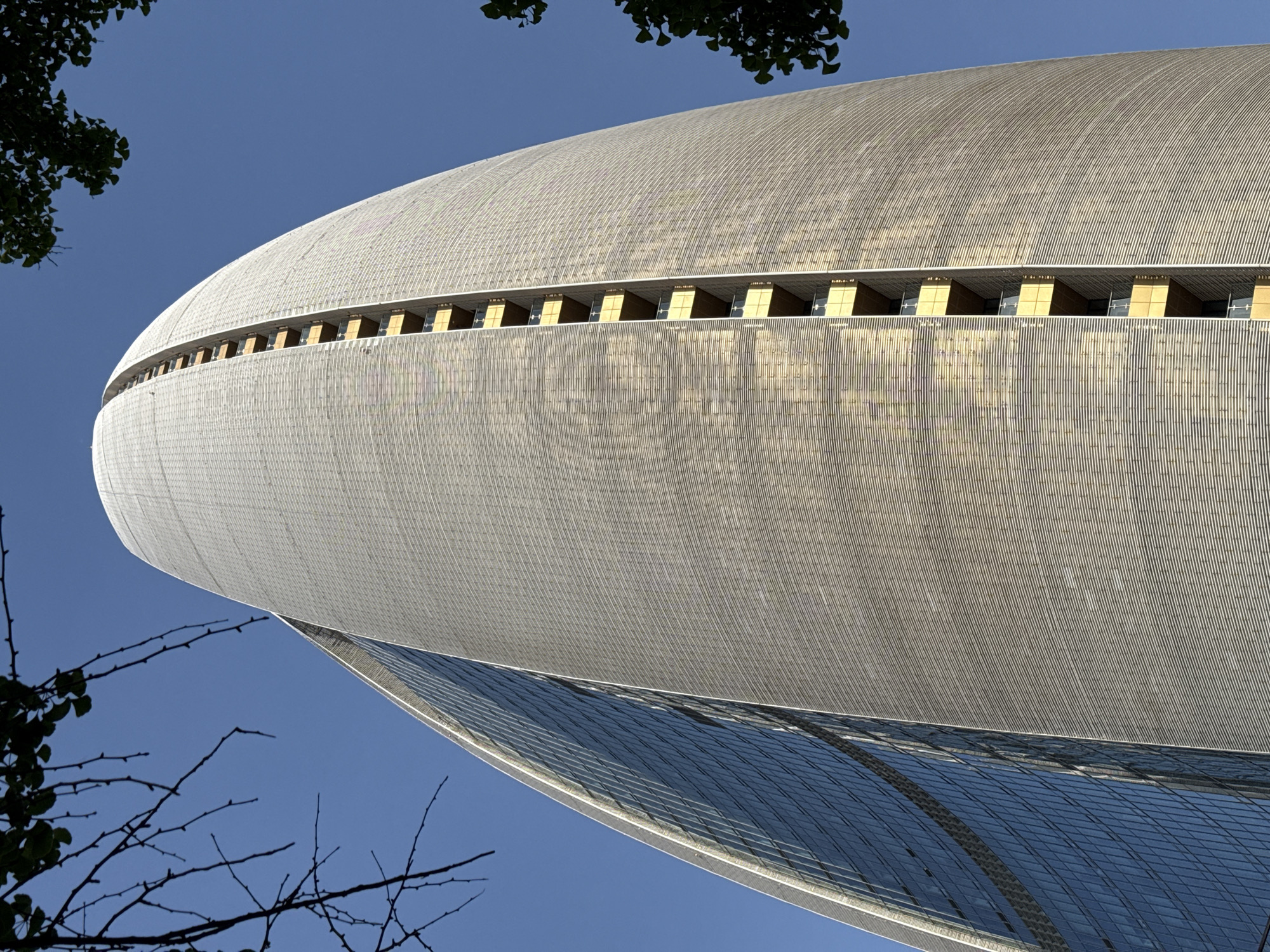 The image shows a low-angle view of the Phoenix International Media Center, located in the Chaoyang District of Beijing, China. The architecture is characterized by its curvilinear form, featuring a metallic exterior with a pattern of vertical lines or ridges. A horizontal band of windows runs along the building's curvature. The lower portion of the building appears to be constructed with reflective blue glass. In the foreground, branches and leaves from trees partially obscure the view, suggesting an outdoor setting. The sky in the background is clear and blue.