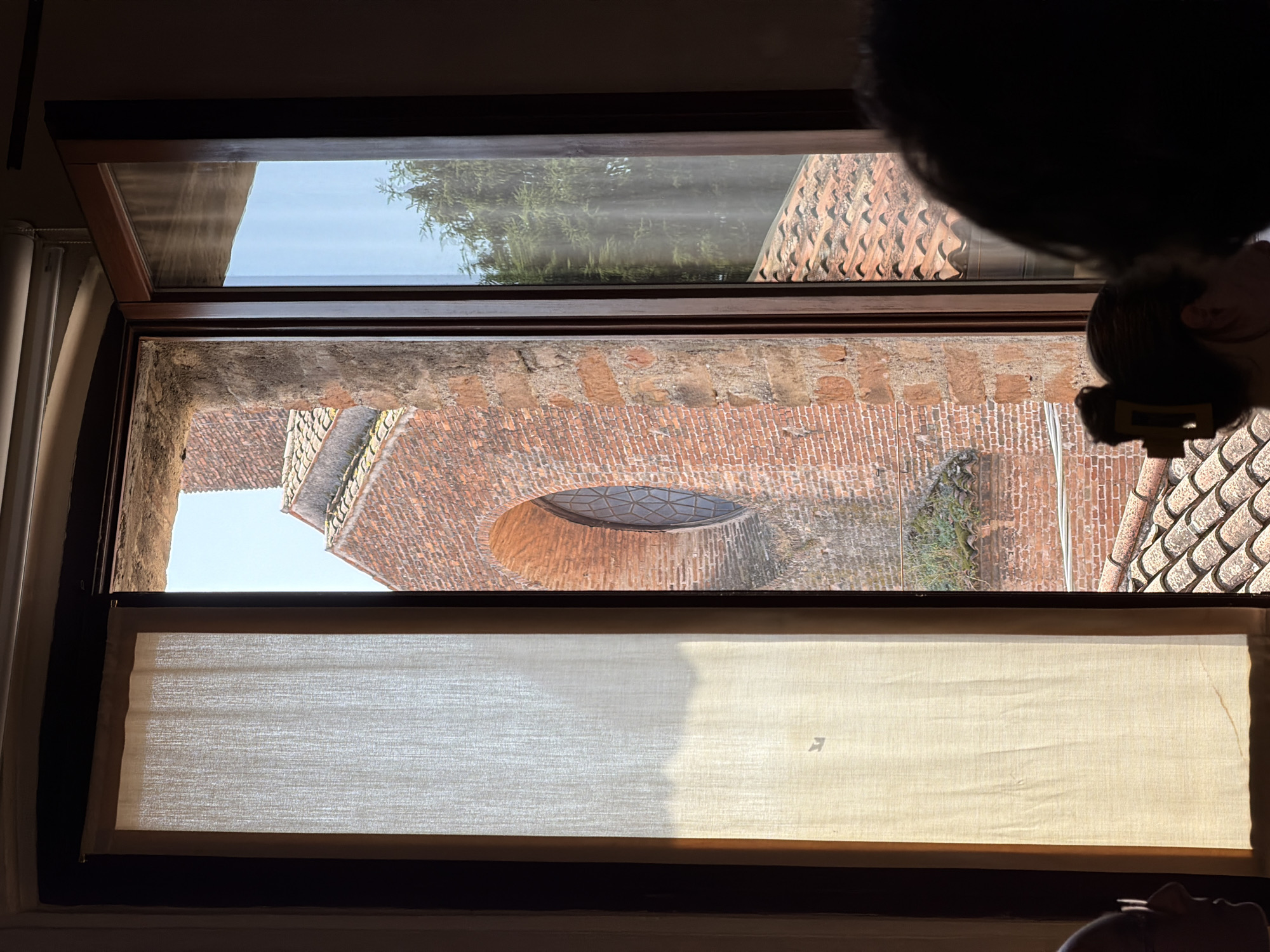A view through a wooden-framed window reveals an exterior scene in Mantua, Italy. The window is partially covered by a light-colored fabric shade, which is partially down. Beyond the window, an aging brick building is visible, with an oval-shaped window towards the building's top. The architecture suggests a historical building or structure, possibly a church. Adjacent to the historical brick building is a sloping tiled roof of a different structure. Outside the window, vegetation covers part of the brickwork. At the top right corner of the frame, there's a dark mass that appears to be part of someone's head, hair, or body. The lighting suggests a sunny day, casting shadows on the fabric shade.