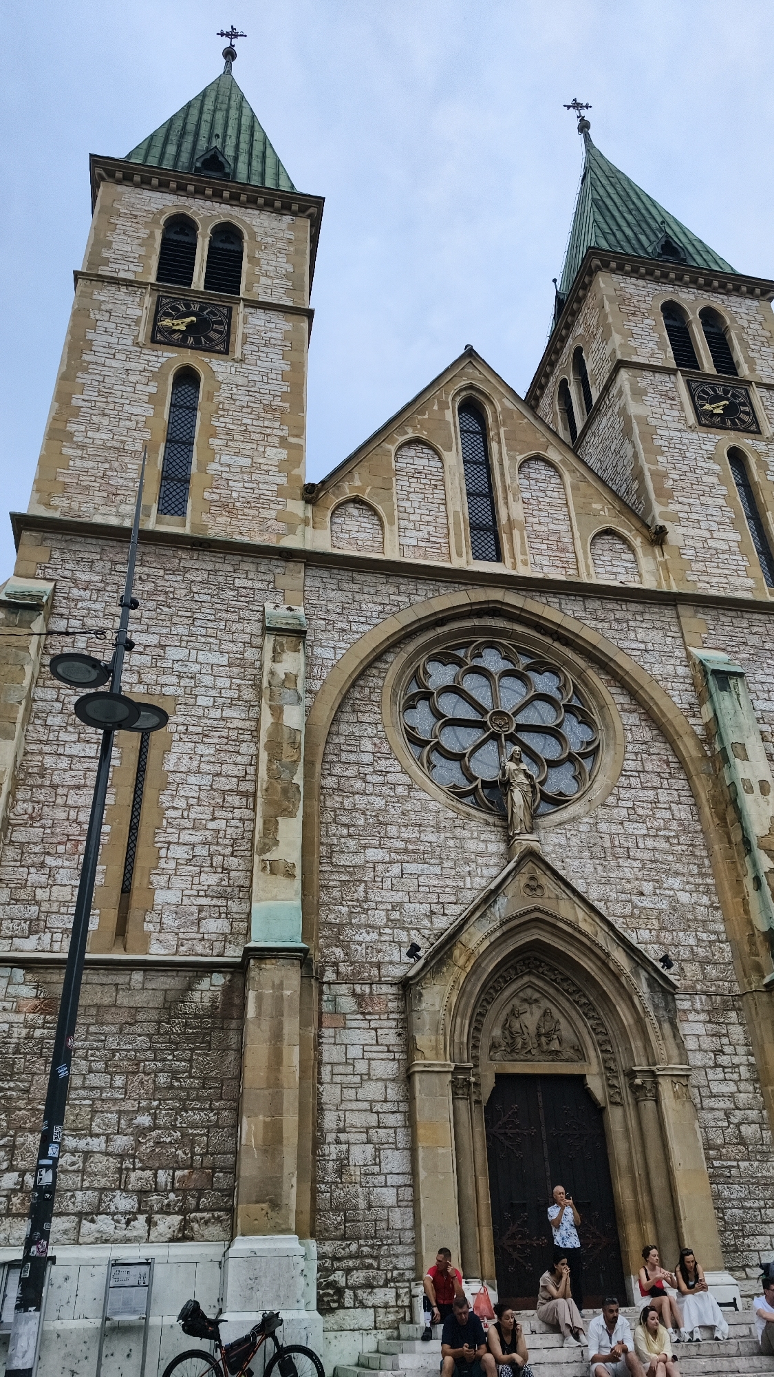 The image shows the facade of the Sacred Heart Cathedral in Sarajevo, Bosnia and Herzegovina. The cathedral features two prominent bell towers with green copper spires, a large rose window above the entrance, and intricate stone masonry. A statue of what appears to be a religious figure stands below the rose window. Several people are seated on the steps leading up to the cathedral entrance. A bicycle is parked to the left of the steps, and a street lamp is visible on the left side of the image. The sky is overcast.