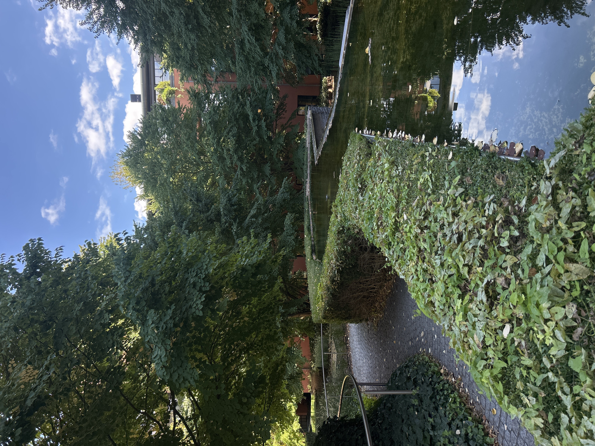 The image depicts a tranquil courtyard with a winding path and a reflective pond, surrounded by lush greenery and a red brick building. A silver railing guides visitors along the paved pathway, which is bordered by manicured hedges and ivy-covered ground. The pond, with its still water, mirrors the blue sky and scattered clouds, as well as the surrounding trees and building. The architecture of the building suggests a modern design with a flat roof and large windows, hinting at a residential or commercial complex. The overall atmosphere is serene and well-maintained, likely a public space designed for relaxation and aesthetic enjoyment within the urban environment. This scene is located in the city of Leuven, Belgium.
