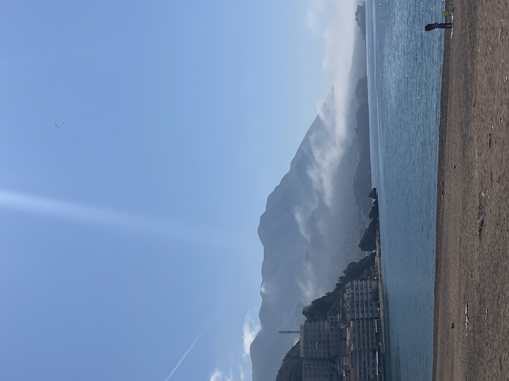 The scene is a coastal landscape, likely near Rafailovići, Montenegro, featuring a beach, water, mountains, and buildings. The sky is mostly clear blue with some wispy clouds and a contrail from a plane. In the distance, a large, hazy mountain range is partially obscured by mist or clouds. The base of the mountains transitions to a built-up area with several tall buildings, seemingly residential apartments. The water appears to be calm and is a light blue color, reflecting sunlight. A sandy beach stretches along the right side of the frame. A person is standing at the edge of the water. Scattered debris or objects are visible on the sand.