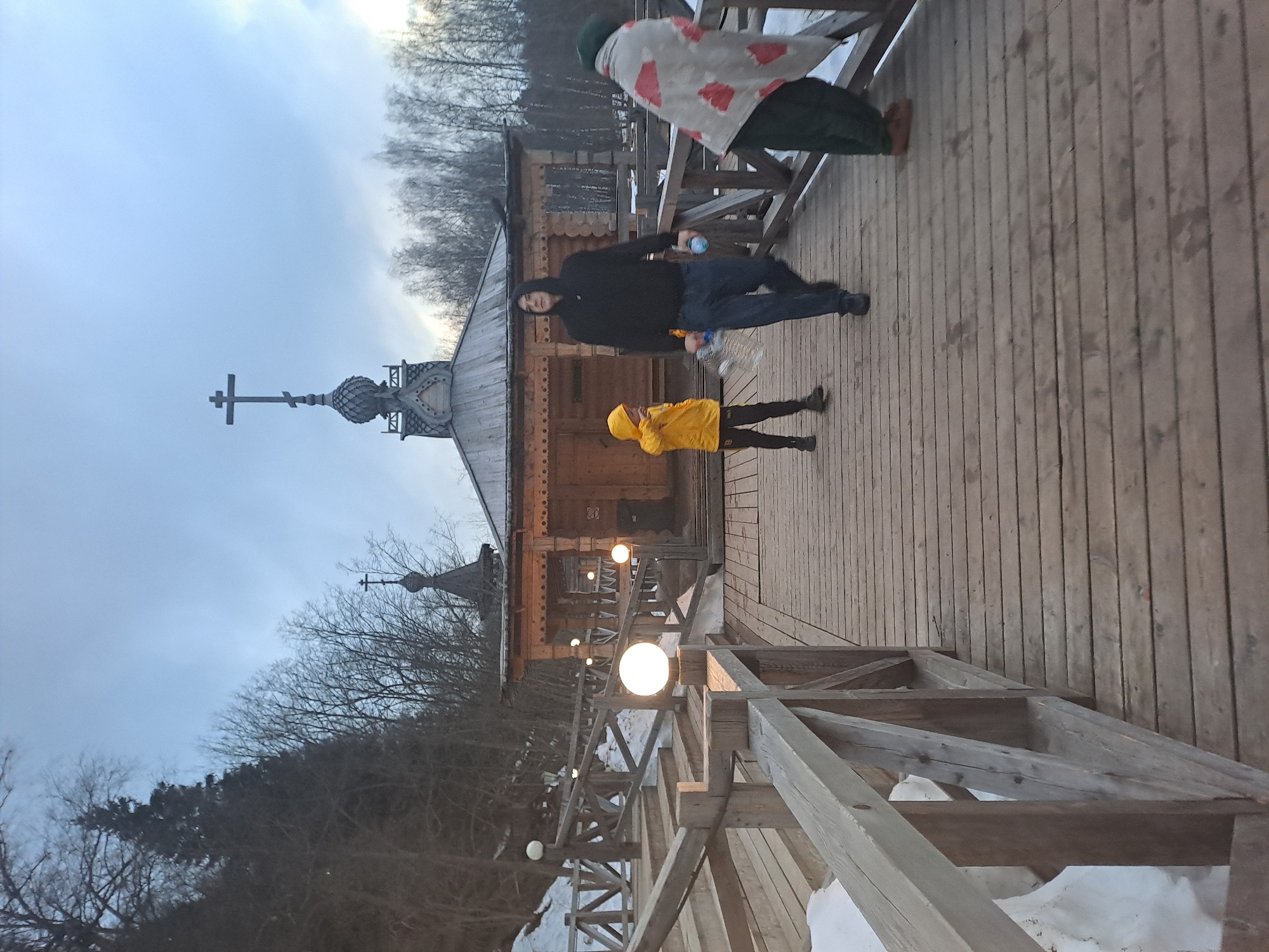 The image shows a wooden church in Vzglyadnevo, Russia, with a bridge leading to it. Three people are on the bridge: one in a green and red blanket, one in a black jacket and jeans, and one in a yellow hooded jacket. The bridge is wooden, and the church is constructed of logs, with a distinct onion dome topped with a cross. Several globe-shaped lights illuminate the bridge. Snow patches are visible near the base of the bridge supports. The sky is overcast.