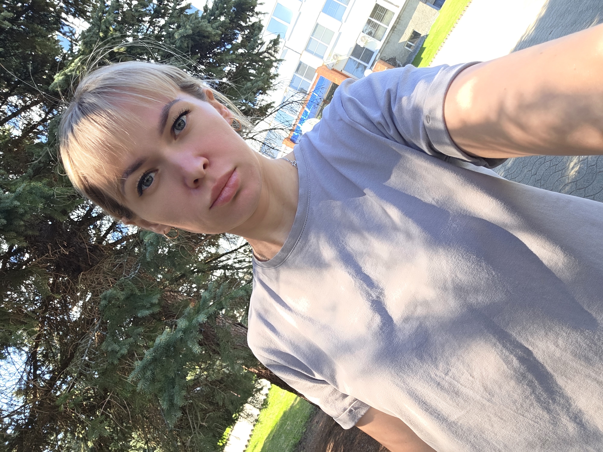 A Caucasian woman with blonde hair and bangs, wearing a gray T-shirt, is taking a selfie. She has light skin, blue eyes, and is looking directly at the camera with a neutral expression. The photo is taken outdoors during the day, with a tree with green needles visible on the left side of the frame. Behind the woman and the tree, there is a multi-story building with white walls and windows. The building appears to be a residential or commercial structure. The ground is covered with grass and a paved area. The specific location is in the city of Chaikovsky, Russia, as per the instructions.