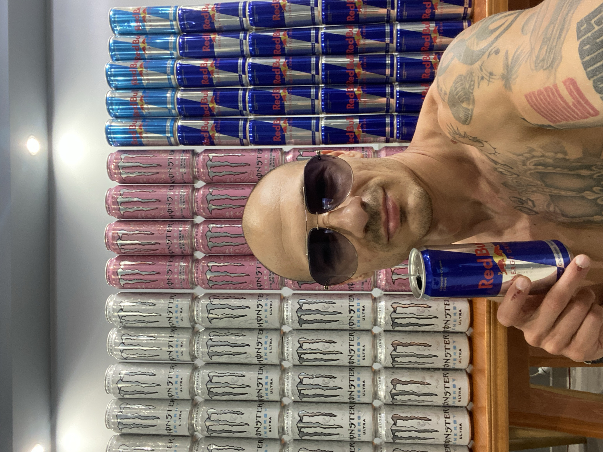 The image shows a man holding an open can of Red Bull, standing in front of a wall stacked with numerous cans of Monster Energy and Red Bull. The man is shirtless and wearing sunglasses. The background consists of rows of silver and pink Monster Energy cans, and blue and silver Red Bull cans, arranged in a structured display. The lighting suggests an indoor environment, possibly a store or a personal collection display. The scene does not provide any specific clues to its location within the city of Pleven, Bulgaria.