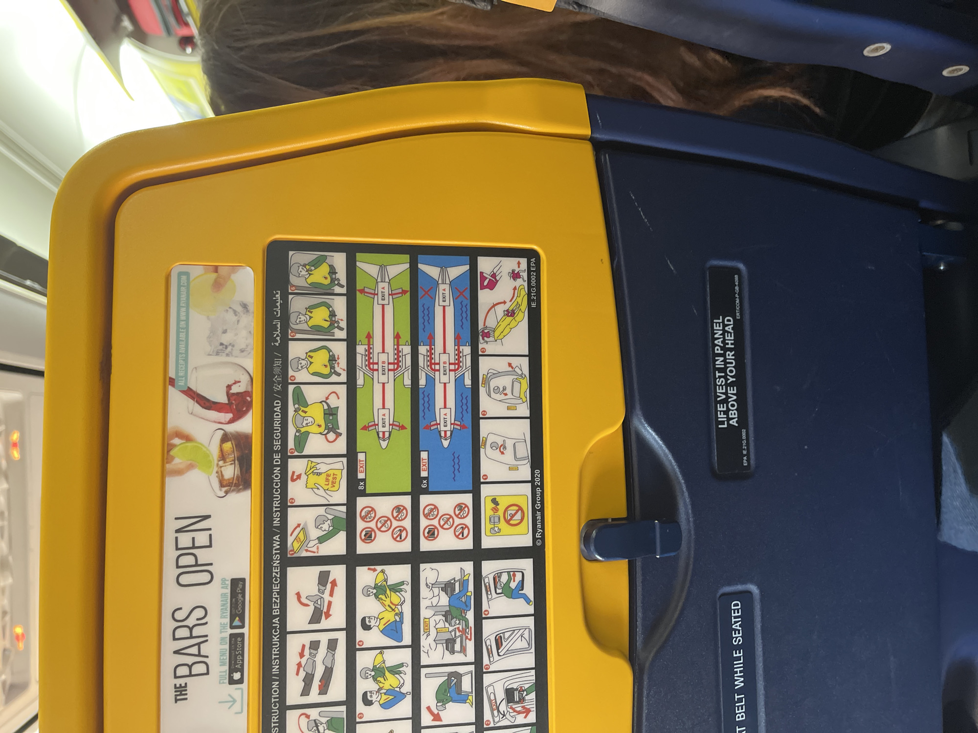 The image shows the back of an airplane seat. It has a yellow folding tray with safety instructions and information about Ryanair's bar service printed on it. Below the tray is the top of a navy-blue airplane seat. Text indicates the location of the life vest. The location, Somma Lombardo, Italy, is not apparent from the image.