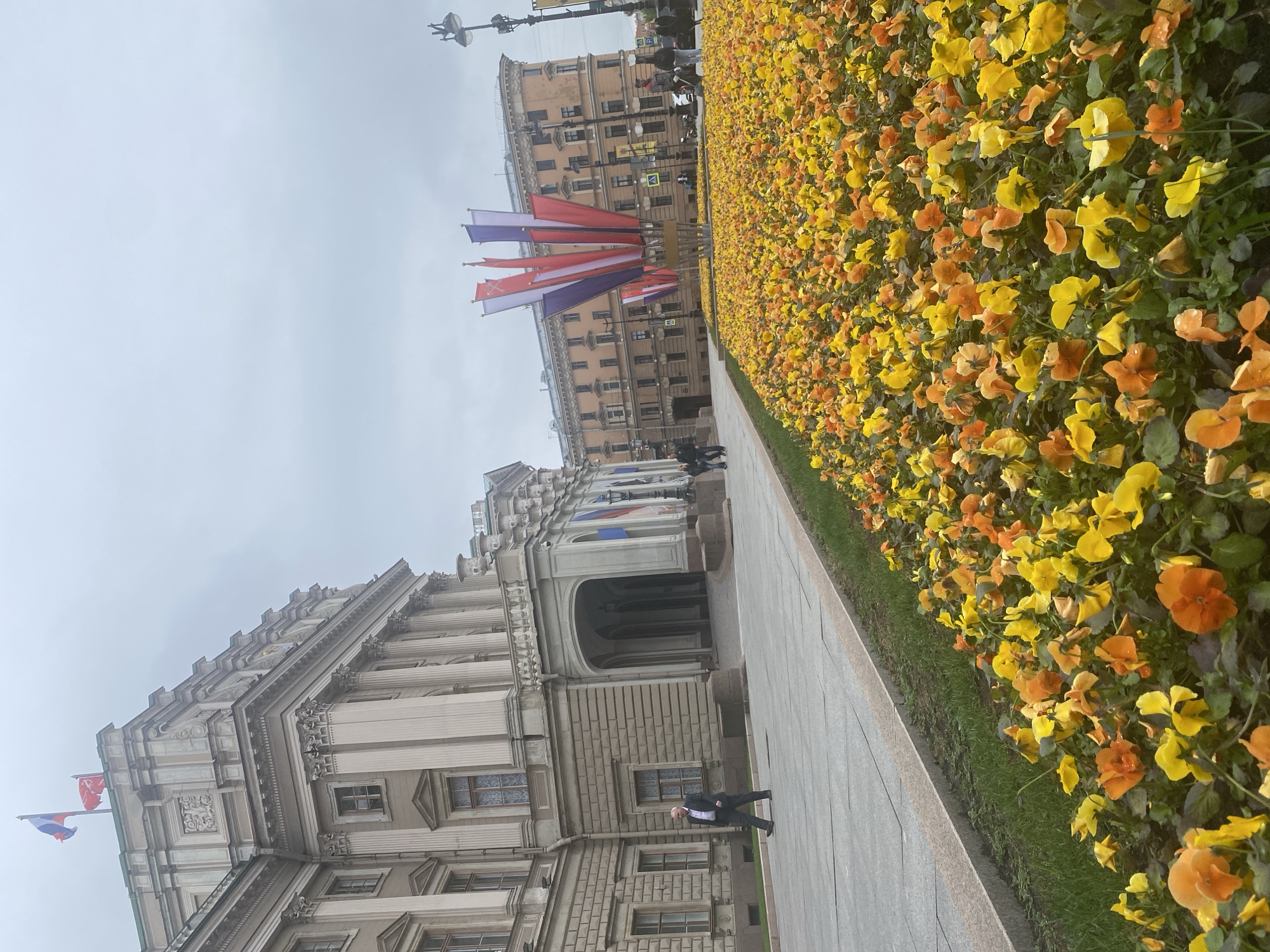 The image depicts a view in Saint Petersburg, Russia, likely near Palace Square. In the foreground is a stone building with classical architectural elements, including columns, windows, and decorative carvings. The building has a Russian flag on its roof. Adjacent to the building is a sidewalk, on which a person dressed in a suit is walking. To the right of the sidewalk is a flower bed filled with yellow and orange pansies. In the background, there is another building and flag-like decorations strung up. The sky is overcast, suggesting a cloudy day.