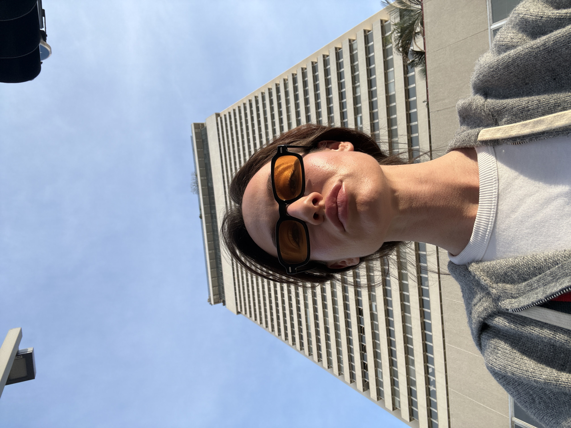 A low-angle, eye-level photograph captures a person wearing sunglasses, with a tall building in the background under a blue sky. The person is wearing a white shirt underneath a gray hooded sweatshirt. The building is a multi-story structure with rows of windows. A traffic light is visible in the upper left corner. The location is likely in Tel Aviv, Israel.