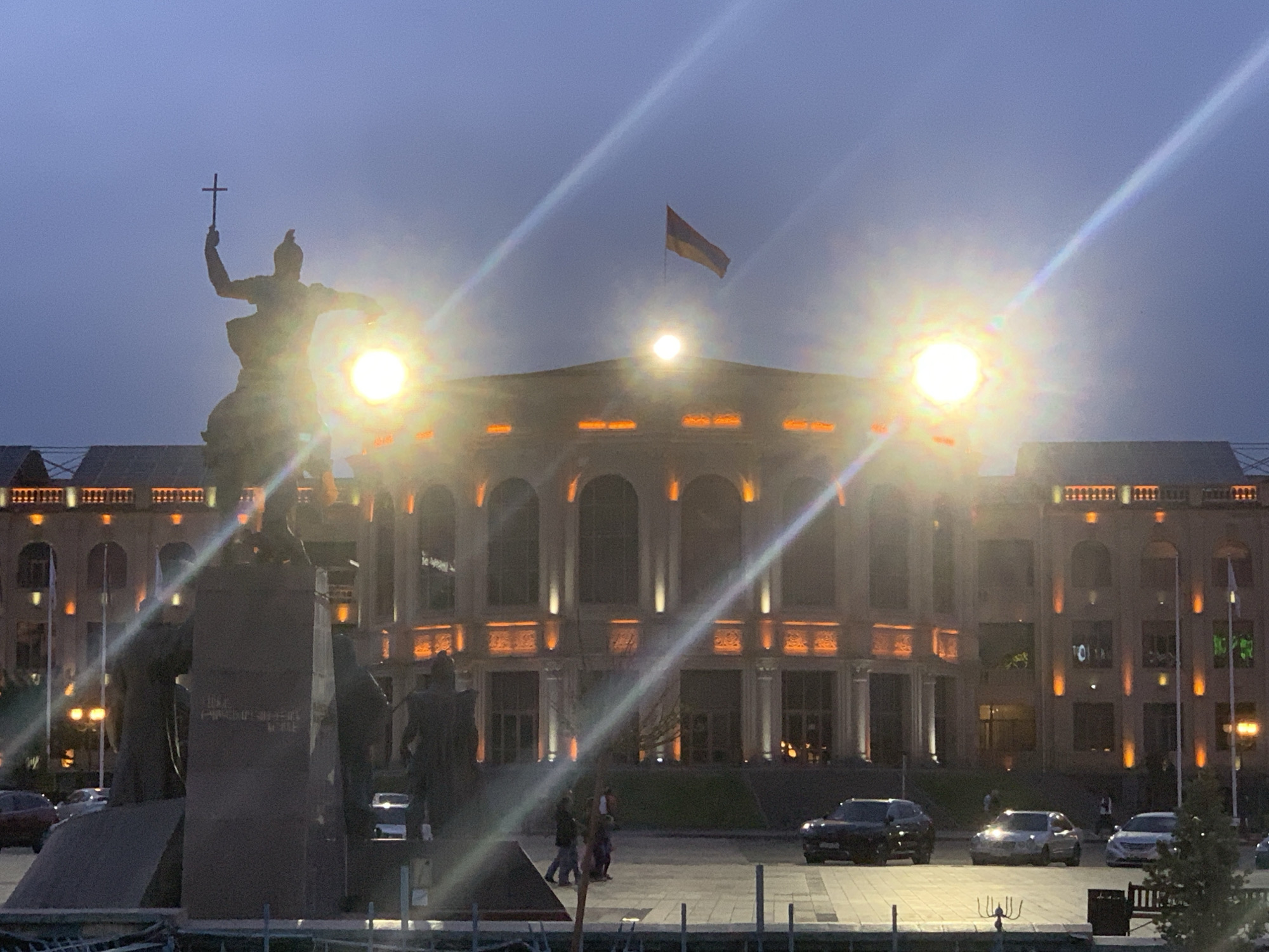 The image depicts Vardanants Square in Gyumri, Armenia, at dusk or early night. In the foreground and midground, a large equestrian statue of Vardan Mamikonian stands on a multi-part stone pedestal. The central figure is on horseback, holding a Latin cross aloft in its raised right hand. Several smaller, dark figures are discernible around the base of the monument.
Behind the statue, the Gyumri City Hall, a multi-story building with classical architectural elements, dominates the background. The building features numerous arched windows and is illuminated by warm, orange-toned accent lights highlighting its architectural details. On the building's roofline, towards the right, the Armenian flag (red, blue, and orange horizontal stripes) is visible, fluttering from a flagpole.
Multiple bright artificial light sources, positioned on the building's roofline and behind the statue, create significant lens flares and starburst effects that diagonally traverse the frame. The sky above is a dark, deep blue, indicative of twilight.
On the paved plaza or street level in the midground, several vehicles are present: a dark SUV on the far left, a light-colored sedan in the center-right, and a white sedan on the far right. A few pedestrians are visible walking across the paved area, appearing as dark silhouettes. Elements of street furniture, including railings and benches, are present in the lower foreground.