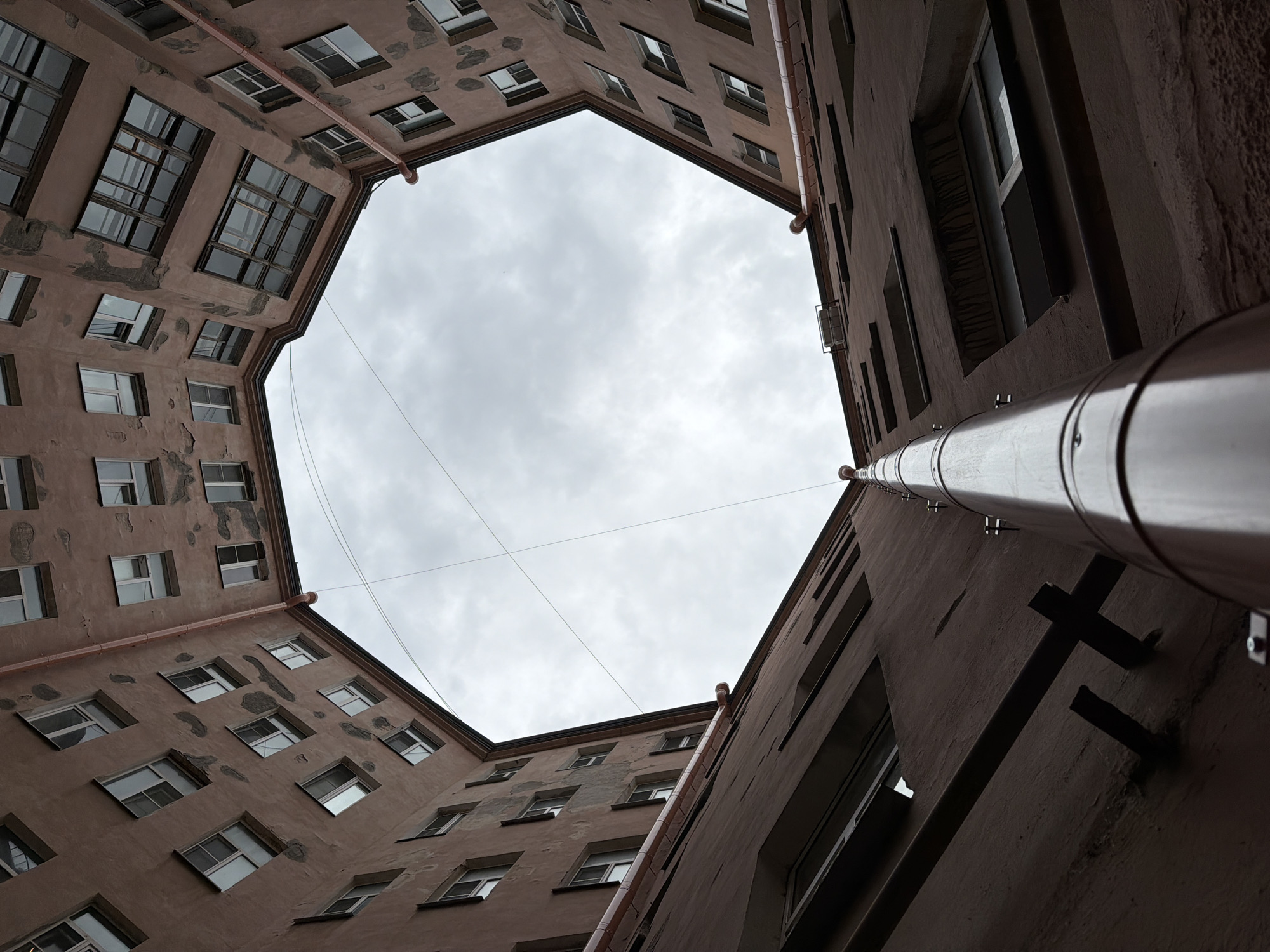 The image shows a view looking upwards into a narrow, octagonal courtyard of an old residential building in Saint Petersburg, Russia. The building's facade is painted a muted, peachy-brown color and shows signs of age, with patches of peeling plaster. Multiple windows, many with dark frames and some with visible panes, are arranged in rows along the walls of the courtyard. A large, shiny, cylindrical downspout runs vertically up the center of the image, attached to the building's wall by metal brackets. A network of thin, dark wires is visible crisscrossing the sky above. The sky itself is overcast with a pale gray, cloudy sky. The perspective of the shot emphasizes the confinement and depth of the courtyard, with the building walls receding upwards and inwards towards the sky.