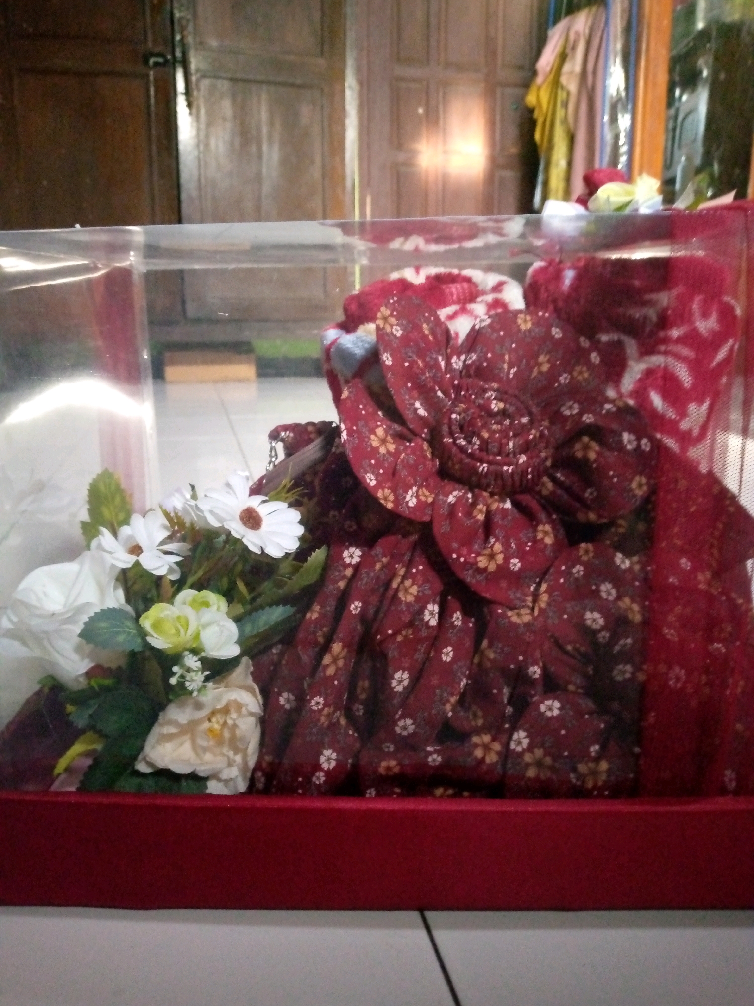 This image captures a close-up view of a gift box containing a red floral fabric, possibly a traditional garment, and artificial flowers. The fabric is predominantly maroon with a delicate pattern of white and yellow small flowers. A prominent, large, multi-layered fabric flower, made from the same maroon floral material, sits atop the folded fabric. In the foreground, a bouquet of artificial flowers is arranged, featuring white daisies with brown centers, pale yellow roses, and smaller white blossoms, all interspersed with green foliage. The gift box itself has a clear plastic lid, and the base is a solid red. The background appears to be an indoor setting, with wooden doors visible, suggesting a home environment in Sumberwono, Indonesia. The lighting suggests an indoor setting, possibly during daylight hours, with some light filtering from the left side.