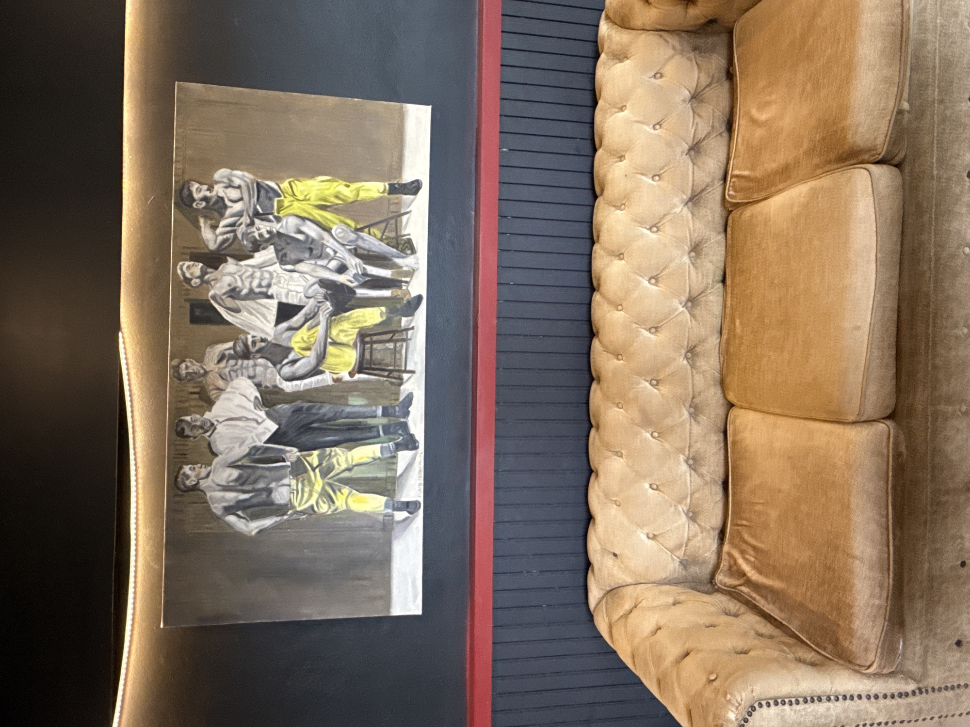A vertical shot shows a room with a painting and a couch. The painting is on a black wall and shows a row of men in yellow pants. Next to the painting is a red strip and then a black wall with horizontal stripes. To the right of the black wall is a light brown tufted couch with three visible seat cushions. The shot is taken from an overhead angle. The location is บ้านบางรักษ์, Thailand.