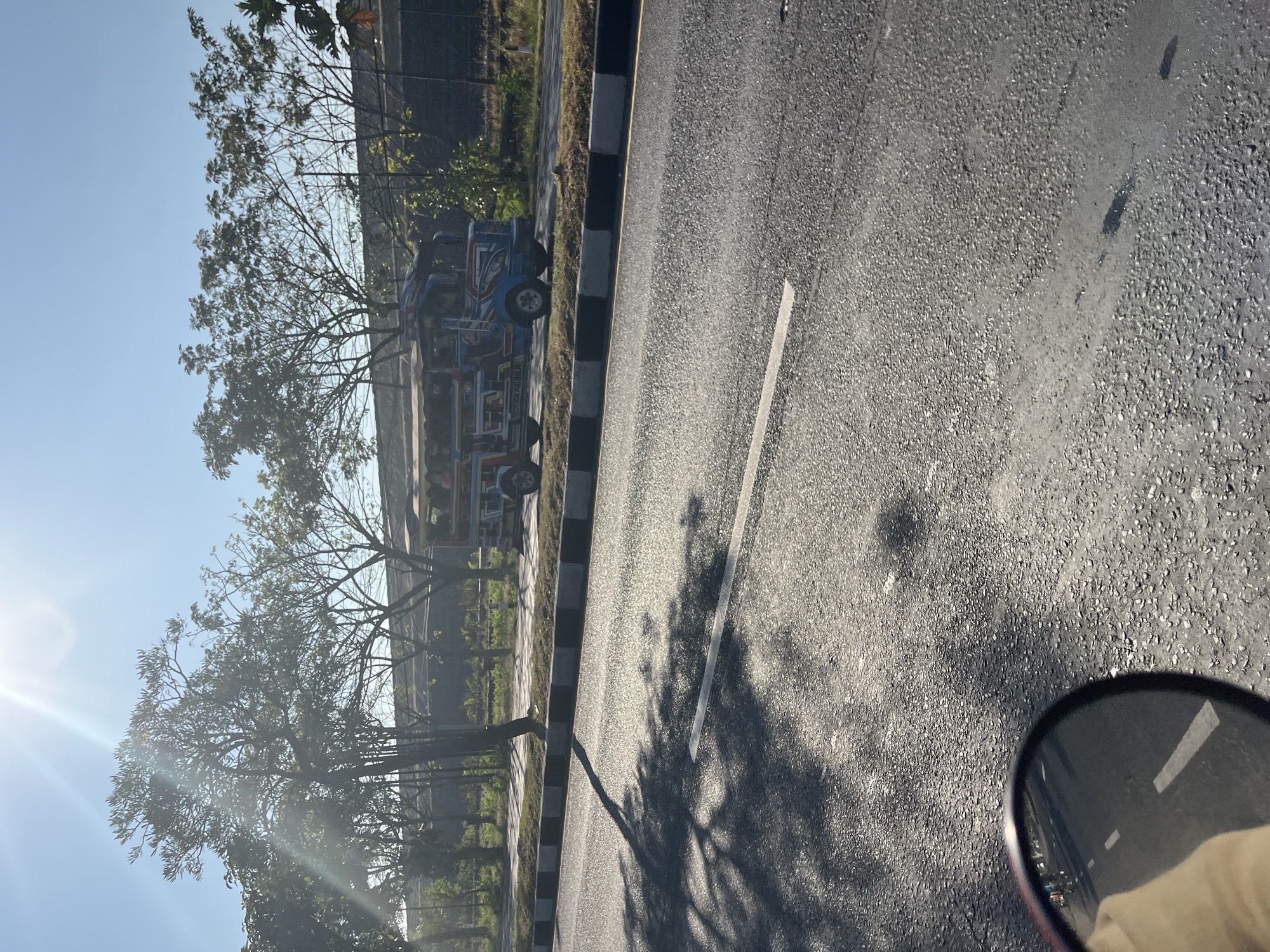 A bright, sunlit scene unfolds on a road in Siring, Indonesia, with a vibrantly decorated jeepney as the central focus. The jeepney, painted in shades of blue, red, and yellow with intricate designs, is stationary on the side of the road. Trees with sparse foliage line the road, casting long shadows on the asphalt. The road itself is divided by white lines, and a black and white striped curb separates it from a grassy area. In the foreground, a portion of a motorcycle's rearview mirror reflects the road and a person's hand. The sky is a clear, pale blue, with the sun's intense glare creating a lens flare effect in the upper left corner. The overall impression is one of a warm, sunny day on a typical Indonesian road.