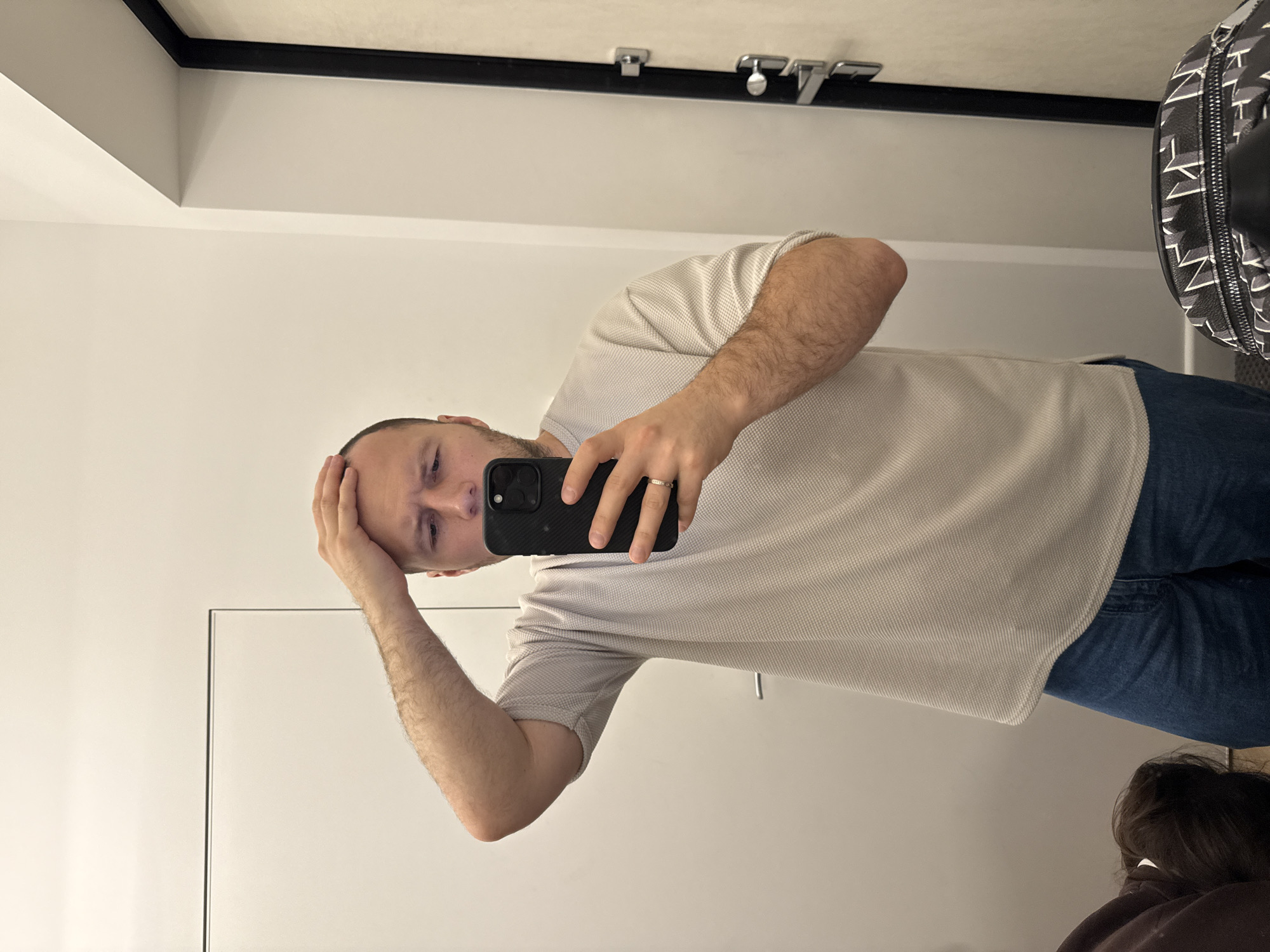 A man is taking a selfie in what appears to be a dressing room. He is wearing a light-colored polo shirt and blue jeans, holding a black smartphone. His left hand is touching his head. The dressing room has plain white walls, a mirror, and a black frame around the top edge. The location in the city of Krasnoyarsk, Russia is unknown.