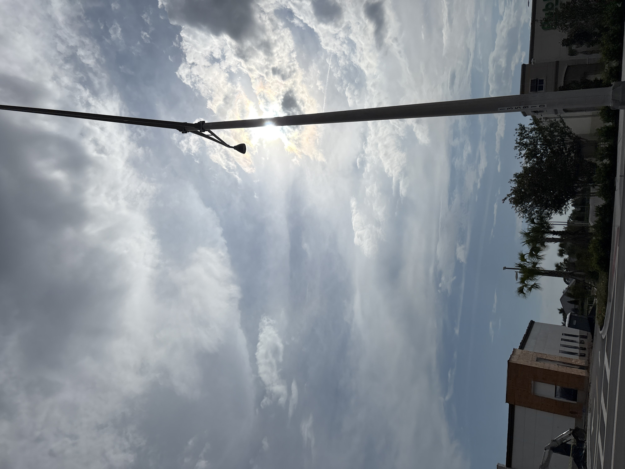 The image shows a street scene in Bradenton, Florida, United States, with a pole in the foreground that has the numbers "135B10" printed vertically on it. The sun is visible behind a cloudy sky, and there are palm trees and buildings in the background. One building appears to be under construction. A portable toilet is located near the construction site. The image is taken from a low angle, looking up at the sky and the pole.