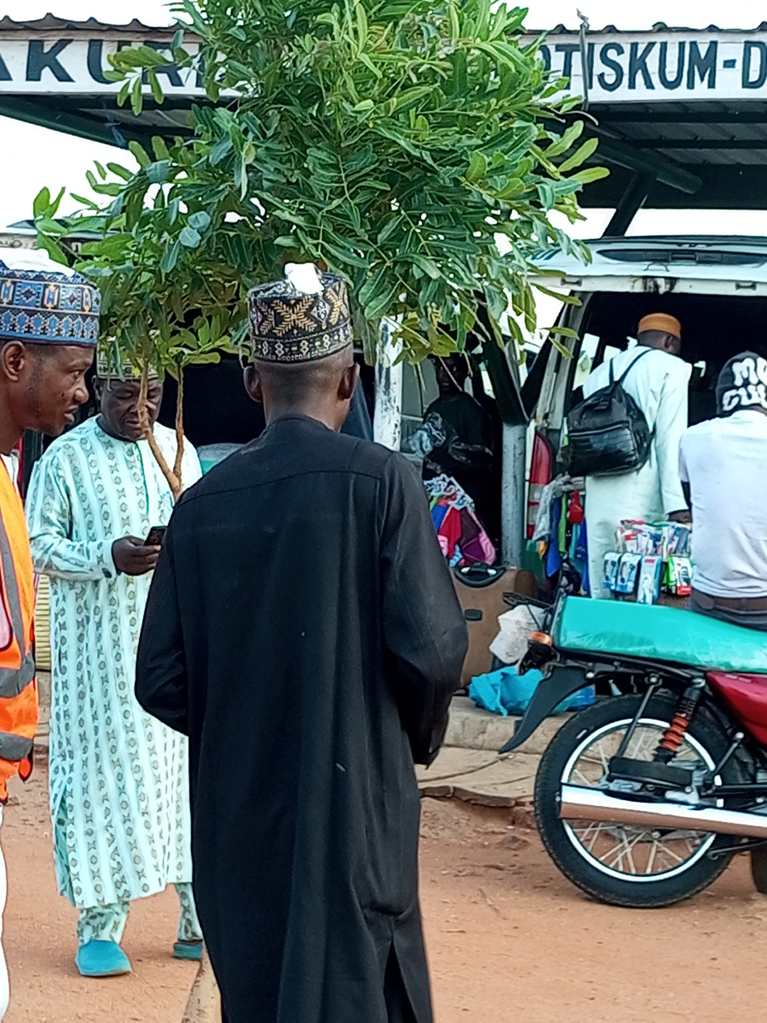 In Gombe, Nigeria, a group of men are gathered around a stall. The stall appears to be set up under a canopy with a sign that reads "AKU GOBIR TISKUM-D". The men are dressed in traditional attire, including caps and robes. One man in the foreground is wearing a black robe and facing away from the camera. To his left, another man in a light blue and white patterned robe is looking at his phone. A third man, partially visible on the far left, wears a reflective orange vest over an orange shirt. Behind them, a van is parked, with its back doors open, displaying various goods for sale, including clothing and packaged items. A motorcycle with a turquoise seat is parked in front of the van. The ground is covered in reddish-brown dirt. The overall scene suggests a busy marketplace or roadside vendor location.