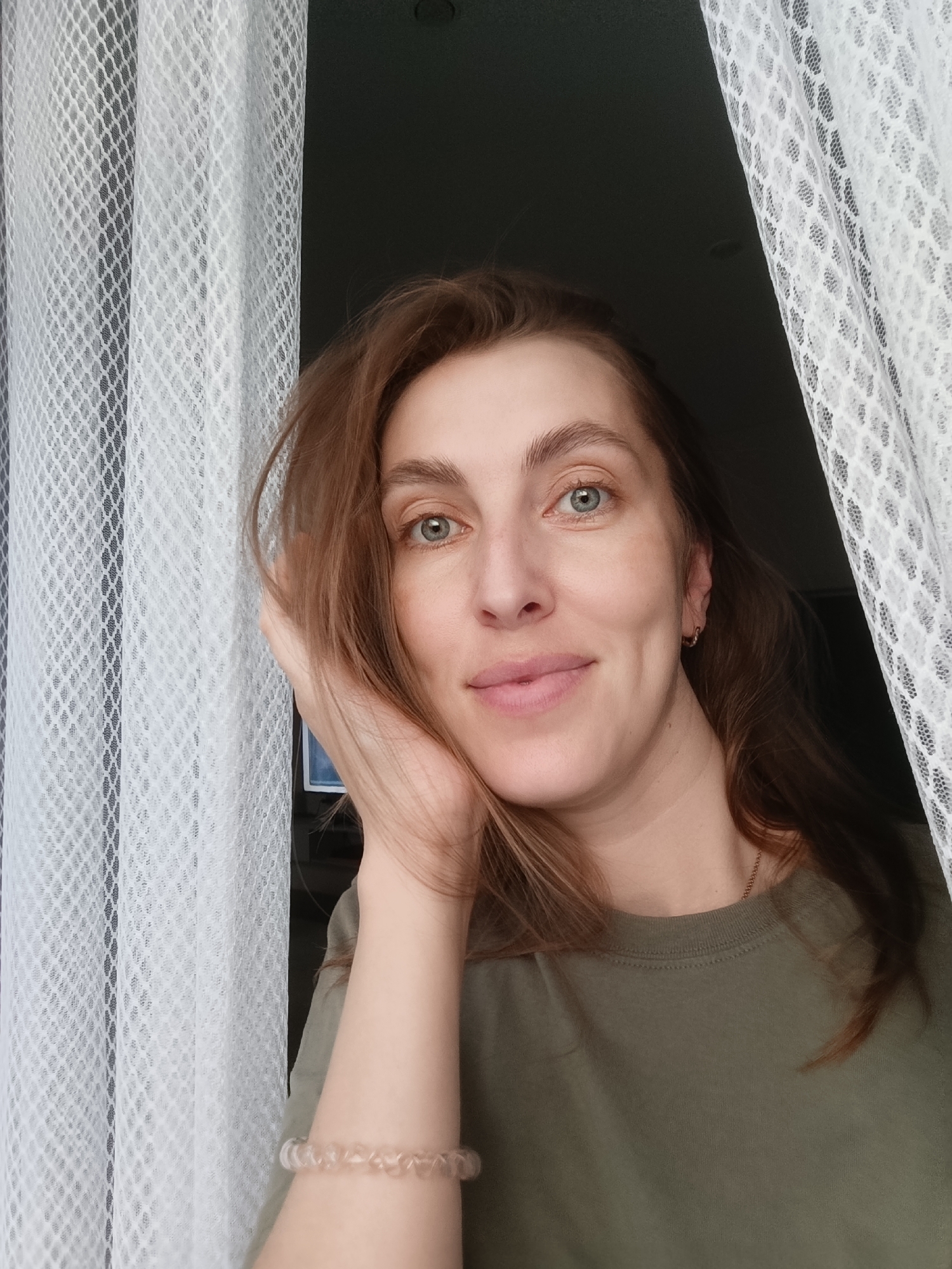 A woman with medium-length brown hair and blue eyes is positioned in the center of the frame, looking directly at the camera with a slight smile. She is resting her right hand on her cheek and her head is tilted slightly to the left. She is wearing a green t-shirt and a light-colored bracelet on her left wrist. She has a subtle dimple on her left cheek and a small hoop earring in her left ear. She is framed by sheer white curtains on both sides, which are slightly out of focus, and a dark background. The lighting suggests an indoor setting, possibly near a window. There is no indication of the city of Chaikovsky, Russia, in the image.