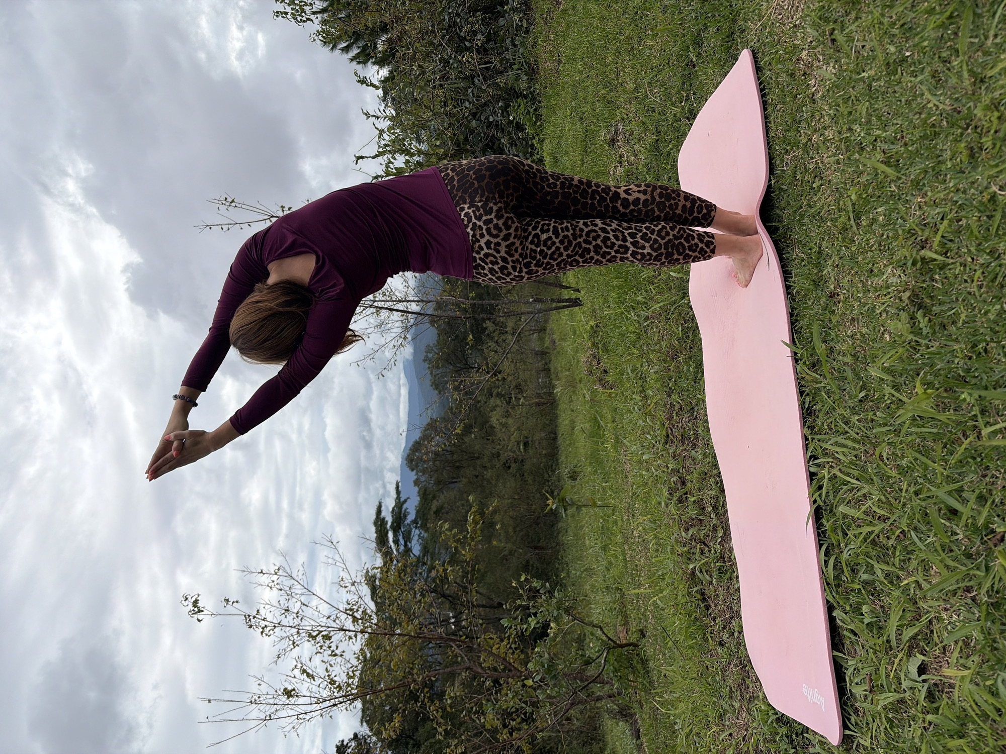 A woman is performing a yoga pose on a pink yoga mat outdoors. She is wearing a long-sleeved plum-colored top and leopard-print leggings. Her arms are raised above her head, palms together, and she is bending to her right, forming an arc with her body. The yoga mat is positioned on a grassy hill. In the background, there is lush green foliage, trees, and a hazy view of mountains in the distance under a cloudy sky. The location is identified as being in the city of Ban Khun Wang, Thailand.