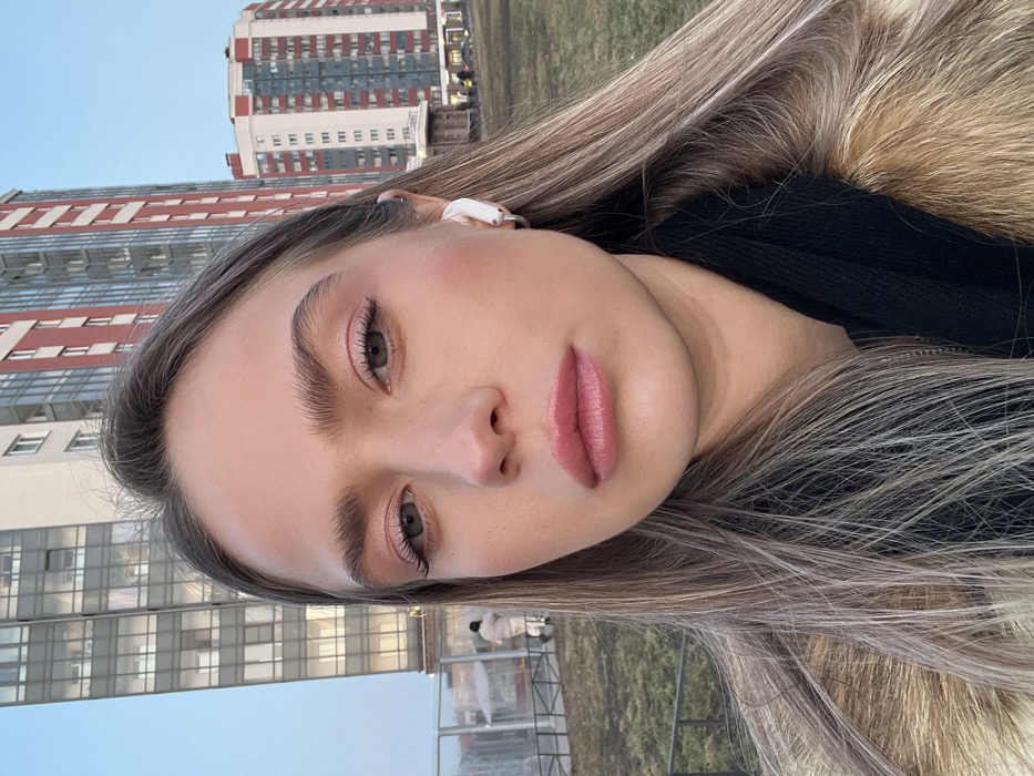 The image displays a medium-close-up selfie of a young adult female, presenting her head and upper torso, taken outdoors in a modern residential area of Saint Petersburg, Russia. The female subject has long, ash-blonde hair, light eyes accentuated with pink eyeshadow and mascara, full pink lips, and well-groomed light brown eyebrows. A white wireless earbud is visible in her left ear, accompanied by a small silver hoop earring. She wears a black garment, possibly a sweater or scarf, beneath a fur-trimmed or fur-lined coat of a light brown or caramel color. Her expression is neutral, with a direct gaze toward the camera.
The background reveals a clear blue sky above a sparse, dry grassy field. Dominating the cityscape are two distinct high-rise residential buildings. The structure on the left is a tall, light-colored edifice featuring numerous reflective glass windows. To the right, another tall building exhibits a distinctive red and white vertical striped pattern on its facade. In the mid-ground on the left, a low metal fence and a partially visible transparent, tent-like structure are present. Several indistinct human figures are visible in the distant background near the buildings, one of whom, dressed in a light-colored coat, walks away from the camera on the left side of the frame. The architectural style of the prominent high-rise buildings identifies the location as a contemporary residential district within Saint Petersburg.