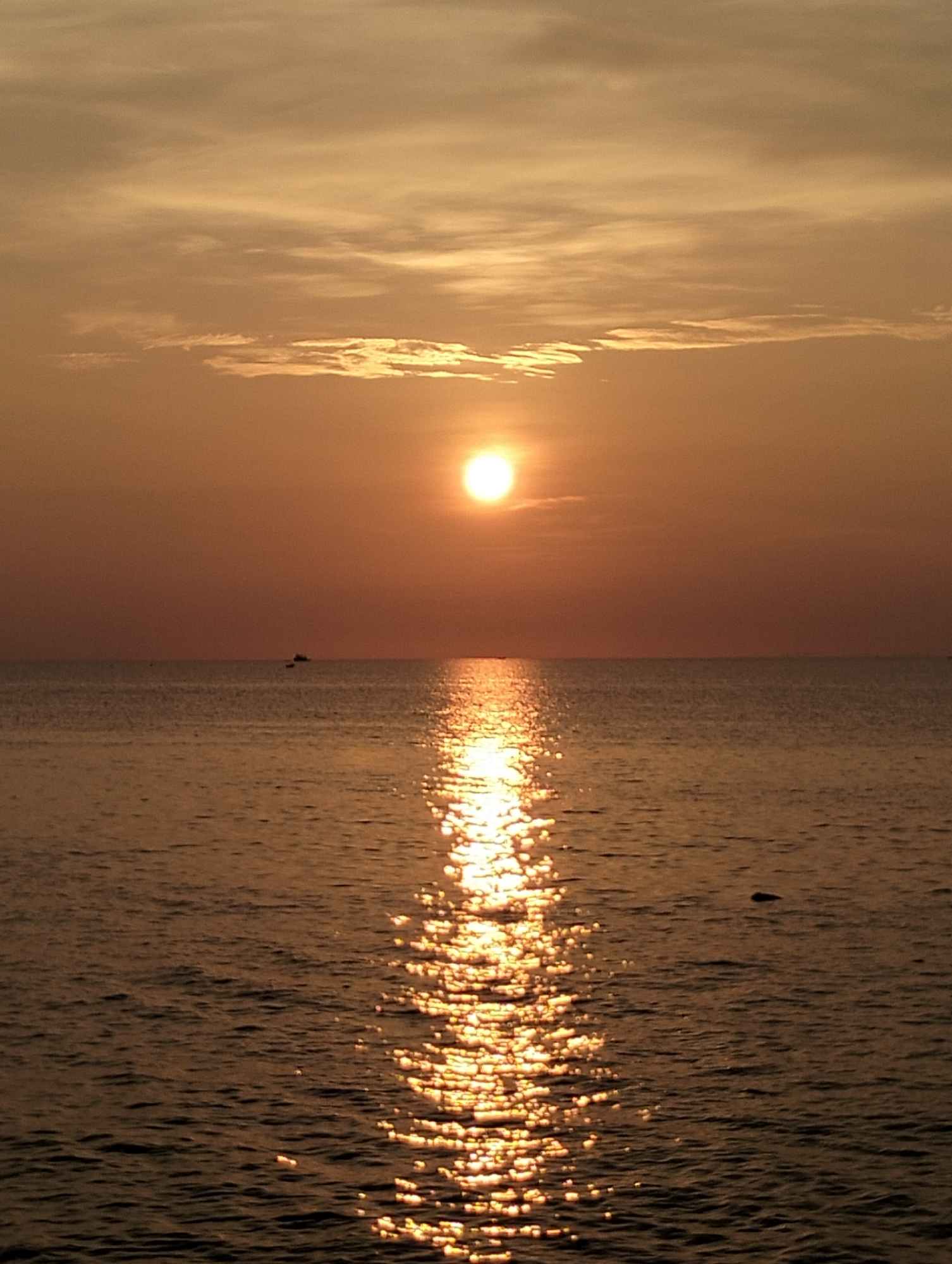 The image shows a sunset over the ocean, likely taken from a location in Lhokseumawe, Indonesia. The sun is positioned high in the sky, obscured by thin clouds. The ocean surface reflects the sunlight, creating a bright, shimmering path towards the viewer. In the distance, a few small boats are visible on the horizon. The sky and water are a warm orange color, typical of sunset lighting.