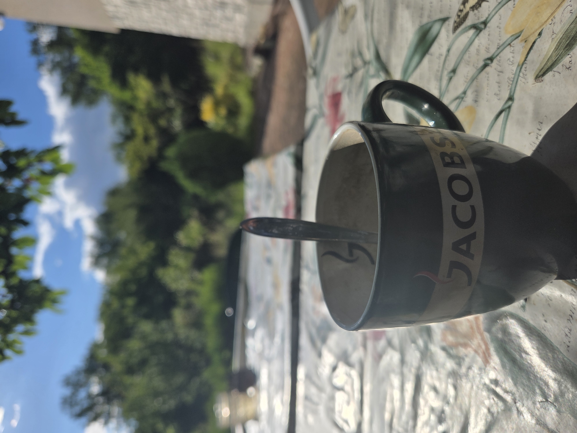 A Jacobs coffee mug sits on a patterned tablecloth outdoors. The mug is dark green with a cream-colored band featuring the Jacobs logo. A spoon is inside the mug. In the background, there are trees, foliage, and a blue sky with white clouds. The scene appears to be a backyard or garden area in Gorna Dikanya, Bulgaria.