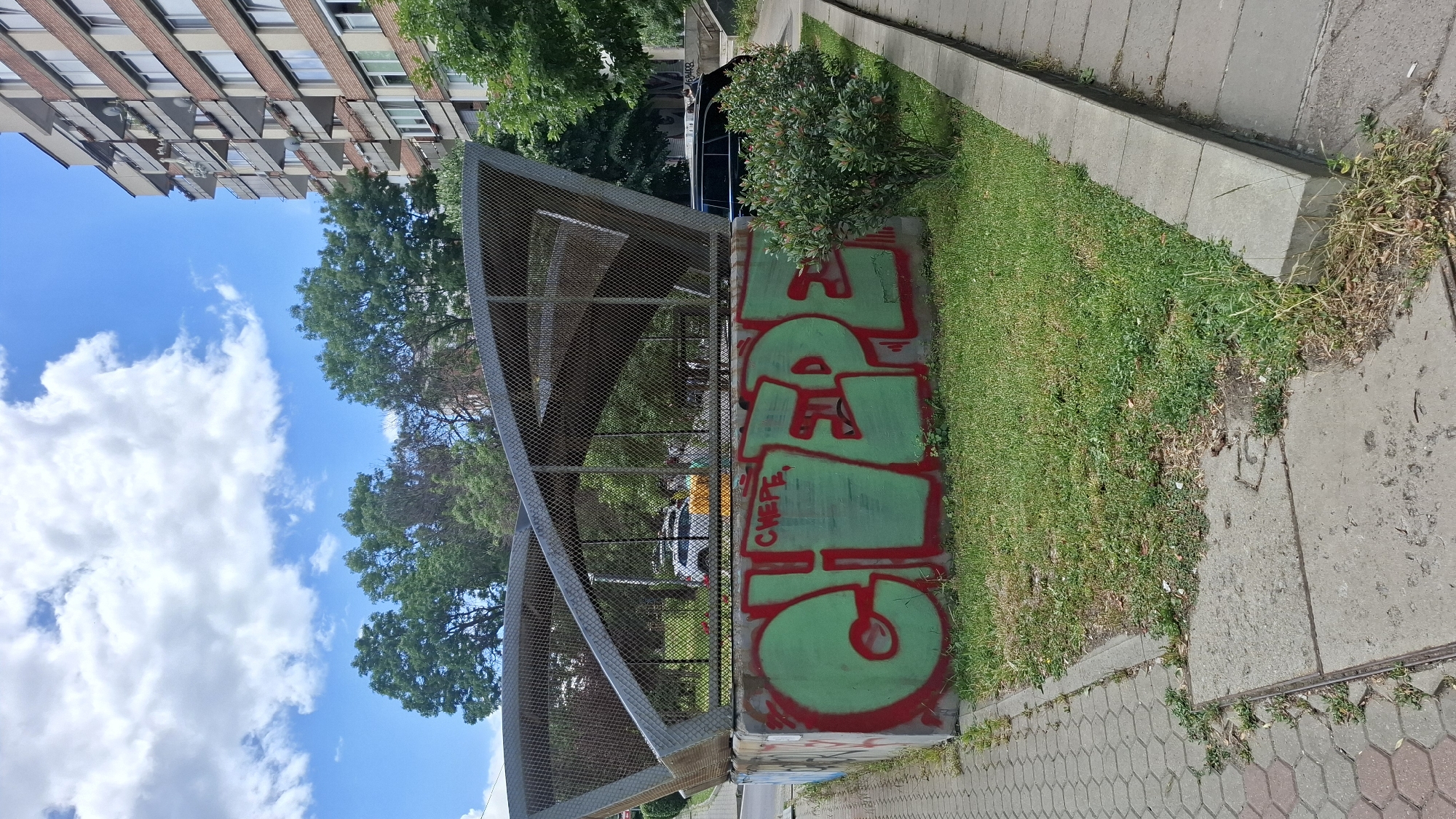 This media file depicts an outdoor urban scene in Sofia, Bulgaria. The main focus is a concrete structure covered in graffiti. The graffiti features large, bold lettering in red outline with a green fill. The word appears to be "CHEPE" or a similar phonetic representation, with "CHEPE" written in smaller red letters underneath the main graffiti. The graffiti is painted on a vertical concrete wall that is part of a raised planter or small bunker. To the left of the graffiti, there is a grey metal fence with a mesh pattern, forming a curved structure that seems to be part of some sort of enclosure or overpass. Behind the fence, trees and foliage are visible, as well as a building with a brick facade and balconies. The sky is blue with white clouds, suggesting it is daytime. To the right of the concrete structure is a paved sidewalk made of grey concrete slabs and interlocking pavers. A strip of green grass runs alongside the sidewalk and the concrete structure, with some patches of dry, overgrown weeds. The scene is viewed from a slightly elevated angle, looking down and to the side. The overall impression is a common urban environment with elements of street art and public infrastructure.