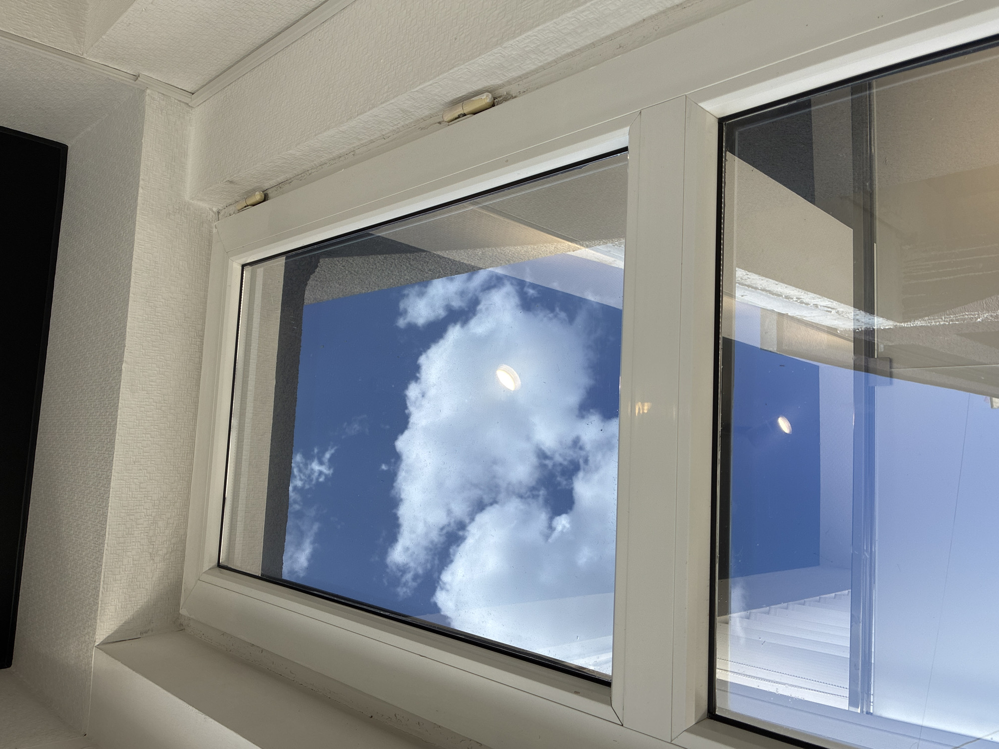 The image shows a close-up view of a white window frame, likely in the city of Khashuri, Georgia. The window reflects a bright blue sky with scattered white clouds. The reflection also captures a circular light fixture. The window itself appears to be divided into two panes, with the top pane showing the sky more prominently and the bottom pane reflecting the sky and some architectural elements, possibly buildings. The interior wall surrounding the window is painted a light color and has a textured wallpaper. A small, beige hinge is visible on the right side of the window frame, attached to the exterior wall. The scene is illuminated by natural daylight.