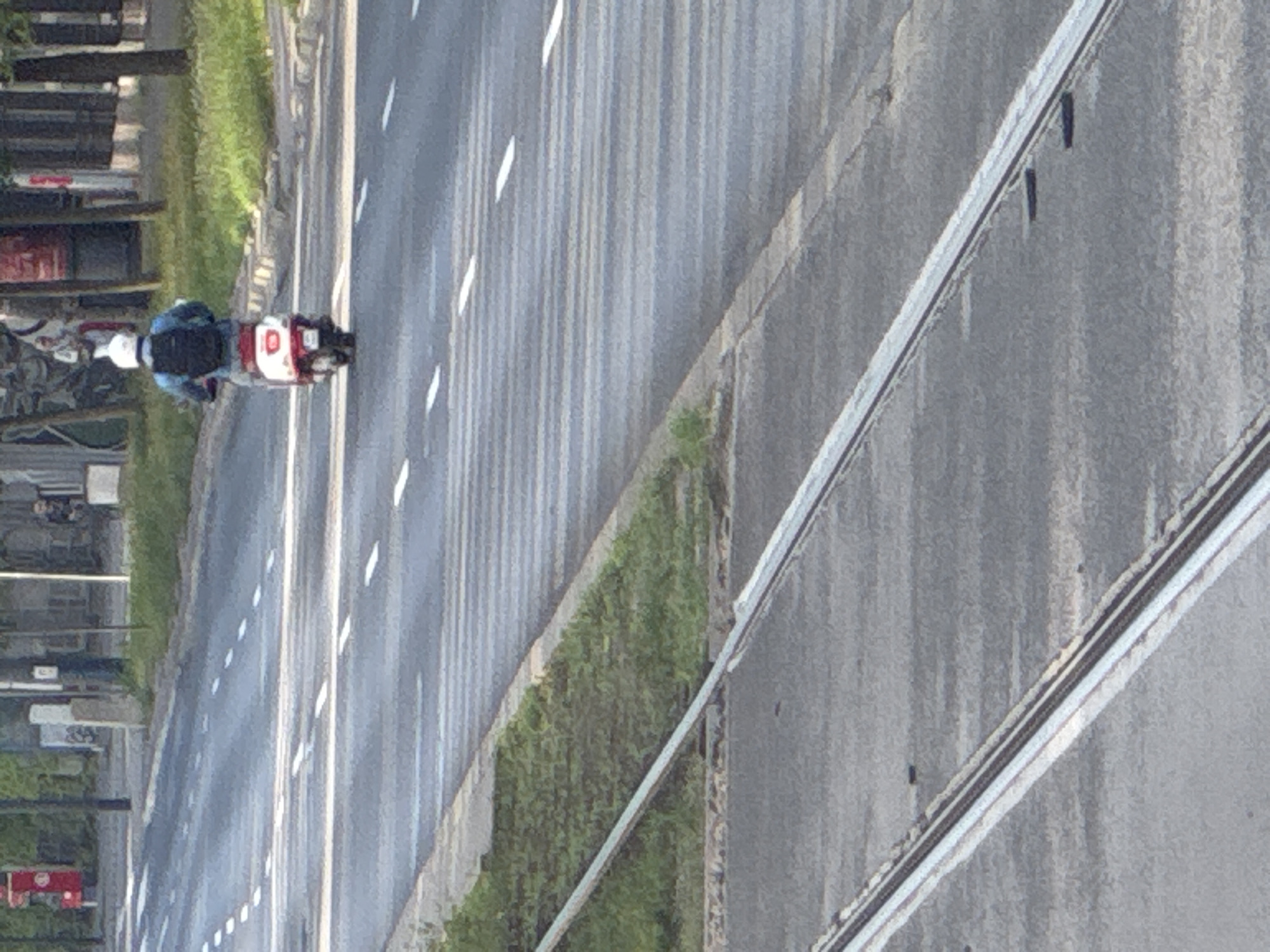 In Warsaw, Poland, a person wearing a white helmet and a blue jacket is riding a red and white scooter on a street. The scooter has a black backpack attached to the back. The street is paved and has white lane markings. There are tram tracks visible in the foreground. The background shows a sidewalk with grass and trees, and buildings in the distance. The scene appears to be during the daytime.
