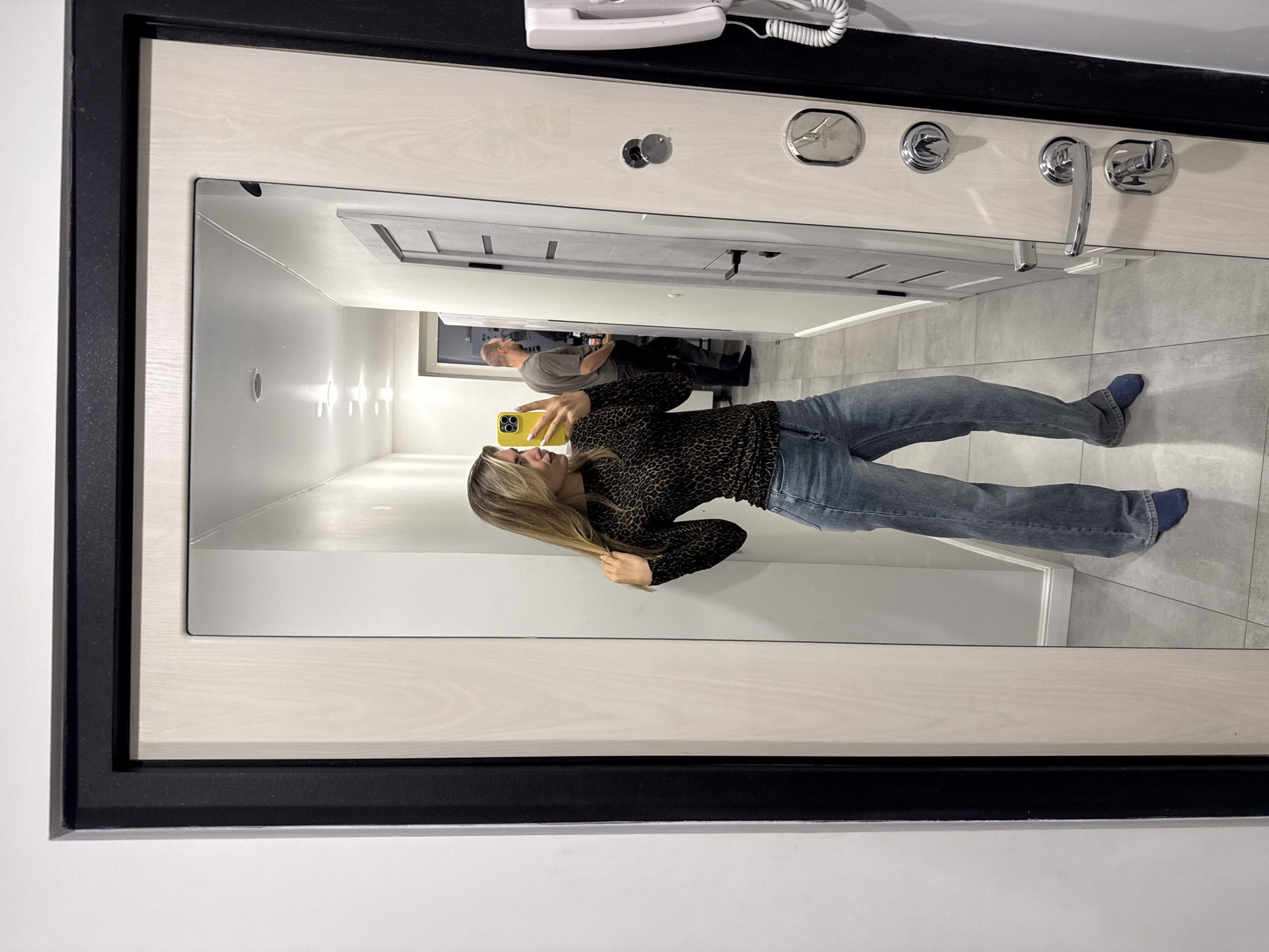 A blonde woman stands in a hallway and takes a mirror selfie. She is wearing a leopard print long-sleeved shirt and blue jeans. The woman is holding a yellow smartphone in her right hand. In the reflection, a man wearing a grey t-shirt is visible in the background. The hallway appears to be an entryway to an apartment or house, with a light-colored door and a white intercom system on the wall to the right of the frame. The floor is tiled in a light gray color.