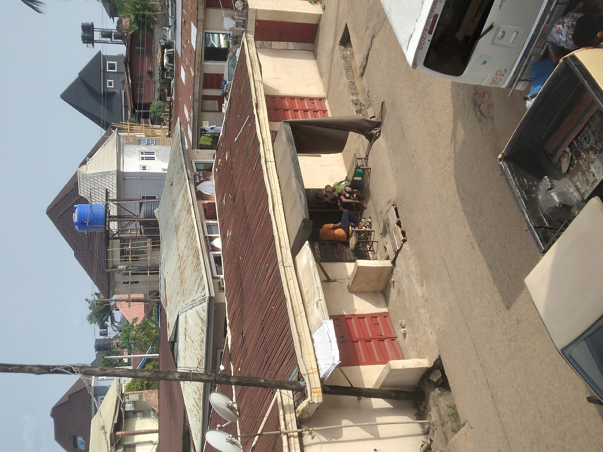 The image shows a street scene in Onitsha, Nigeria. The perspective is from an elevated position, looking down on a narrow street lined with buildings. Buildings have rusty corrugated iron roofs and beige walls. The buildings have red-trimmed doors and windows. Two people sit outside a building. Nearby, a white van and a vehicle with an open bed are parked. An electric pole with satellite dishes is visible in the foreground. Buildings of various sizes and colors are beyond the immediate street scene.