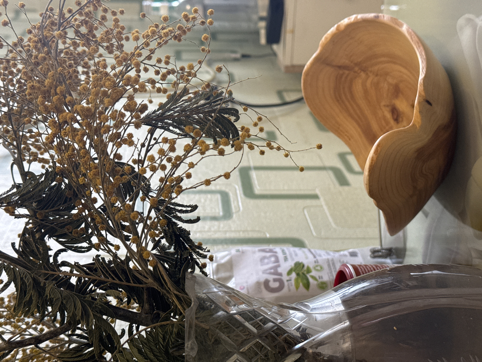 The image shows a still life arrangement on a table in what appears to be an indoor setting. A dried floral arrangement, including yellow pom-pom-like flowers and dark green fern-like foliage, is prominent on the left. Partially visible behind the flowers is a package labeled "GABA TEA SUPPLEMENT." To the right, a rustic wooden bowl or container with natural wood grain is positioned vertically. In the foreground, there's a partially visible clear plastic bag or container with what appears to be more dried material inside. The background features a light green tiled surface with a geometric pattern, and glimpses of a white cabinet or appliance can be seen in the upper right. The location of Ново-Никольское, Russia is not directly verifiable from the content of the image.