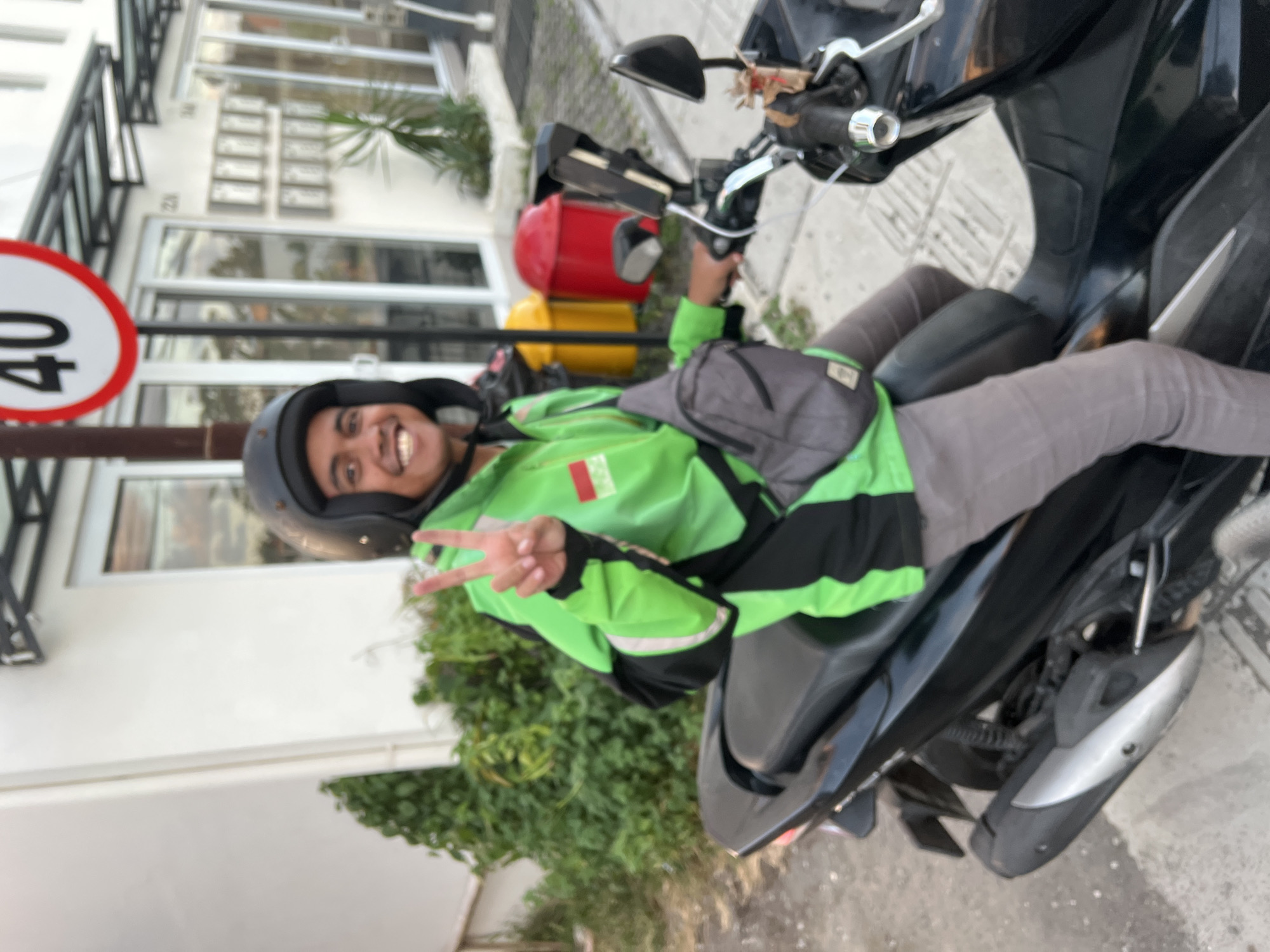 A person wearing a green and black jacket, grey pants, and a black helmet is sitting on a black motorcycle. The person is holding up two fingers in a peace sign. They are wearing a grey bag across their body. The motorcycle has a phone mount attached to the handlebars. In the background, there is a white building with multiple windows and doors. There are also two trash cans, one yellow and one red, next to the building. A speed limit sign with the number 40 is visible above and to the left of the person. The setting appears to be an outdoor urban environment, possibly a street or parking area in Mengwi, Indonesia, as indicated by the architecture and the presence of a speed limit sign. The lighting suggests it is daytime.