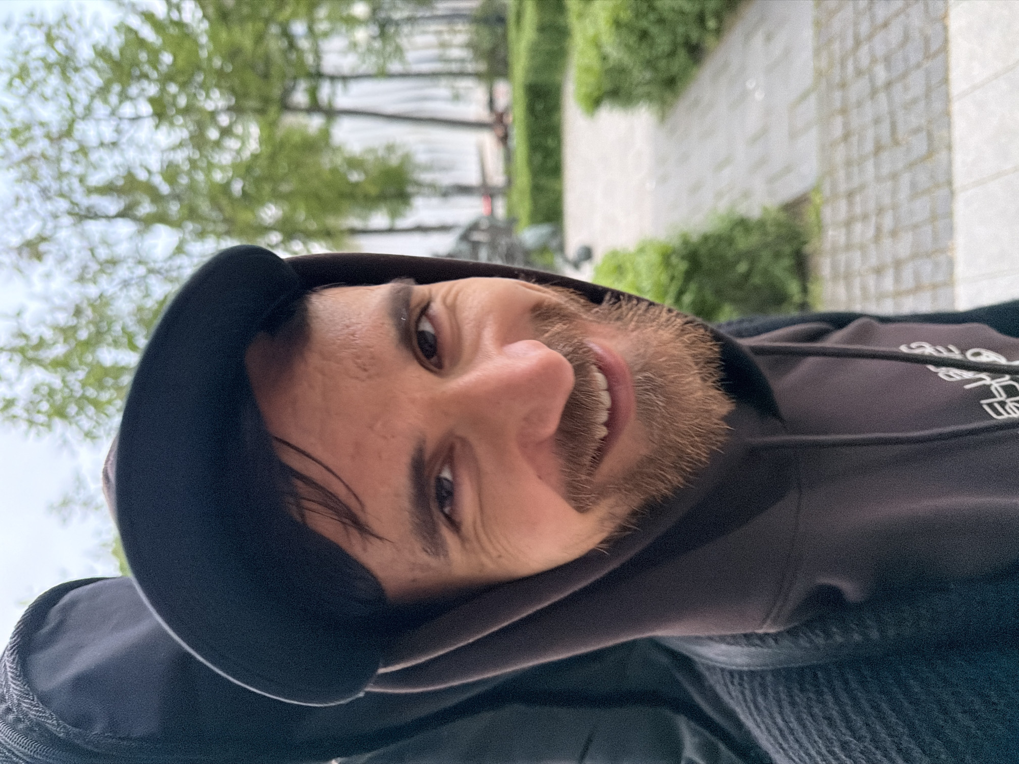 A man with light brown skin, a beard, and dark hair is wearing a dark hoodie and a baseball cap in an outdoor setting. His head is slightly tilted, and he is looking at the camera with a smile. The background shows greenery, trees, a stone-paved surface, and a wall, suggesting an urban environment. Due to the architectural styles of the buildings and the presence of specific landmarks are not visible, it is not possible to definitively confirm that the location is Moscow, Russia.