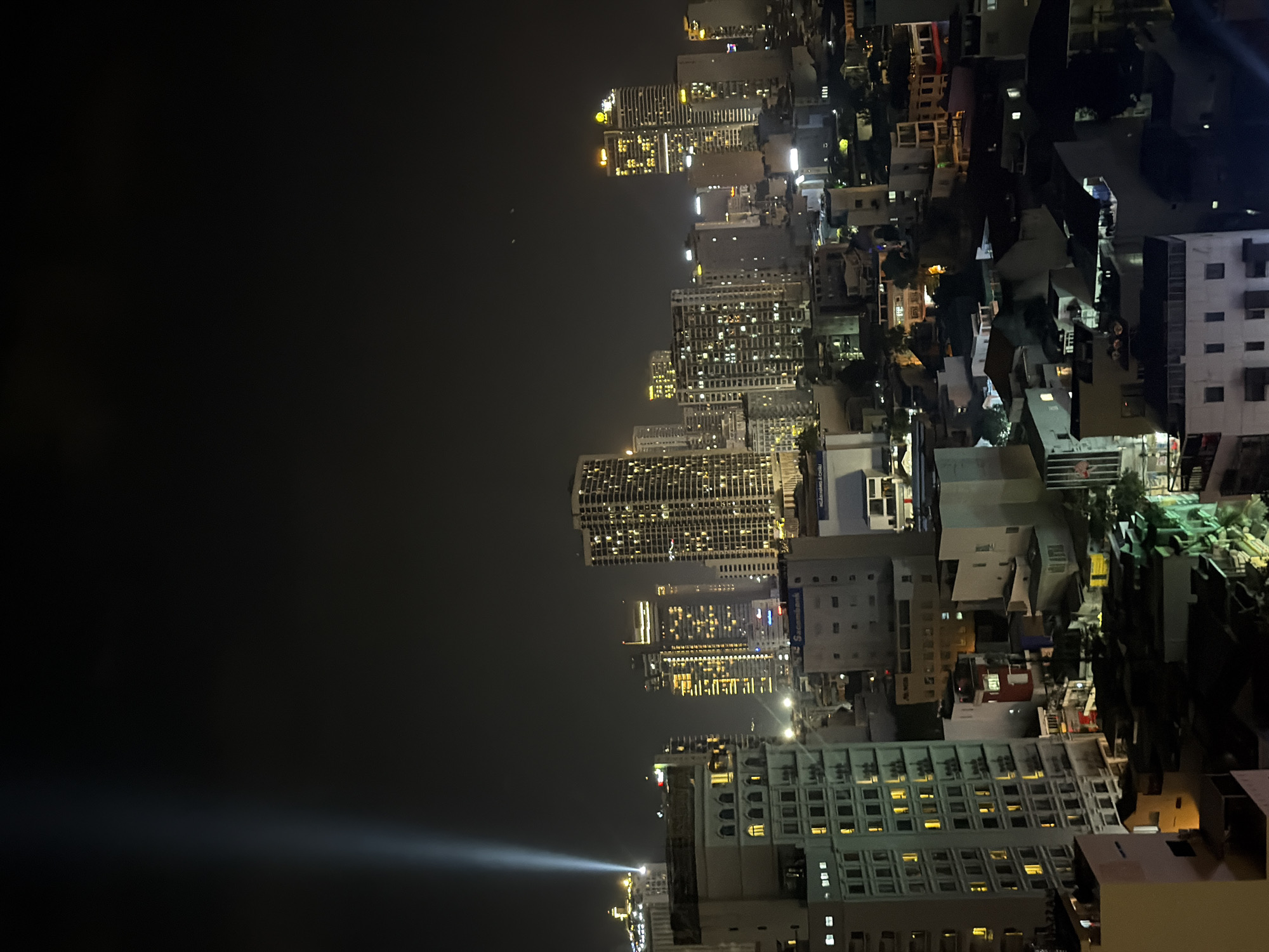 This image presents a nighttime aerial view of an urban landscape identified as Nha Trang Ward, Vietnam. The dark sky occupies the upper half, pierced by a strong, upward-projecting light beam originating from a distant structure on the left side. Two faint, distant light sources are observed in the upper-right quadrant of the sky. The lower half of the image is densely packed with illuminated buildings, indicating active occupancy.
In the foreground, a multi-story building with a light-colored facade is prominent on the left, displaying numerous rectangular windows, many lit with warm yellow light. Its upper levels feature a series of arched windows. Beyond this, a mix of high-rise and lower-rise structures extends into the midground and background. Multiple modern skyscrapers are visible, exhibiting extensive window illumination and distinct architectural lighting, particularly on their upper sections, with some appearing golden.
Identifiable commercial signage includes "Sacombank" visible on a mid-ground building, and a partially obscured sign suggesting "NGAN HANG & CHIU" on another building to its right. The mid- and lower-ground areas are filled with a compact arrangement of smaller, residential or commercial buildings with varying rooflines, many showing illuminated windows. No individuals, specific actions, or direct interactions are discernible within the frame. The scene portrays a bustling city at night, defined by artificial light against a dark sky.