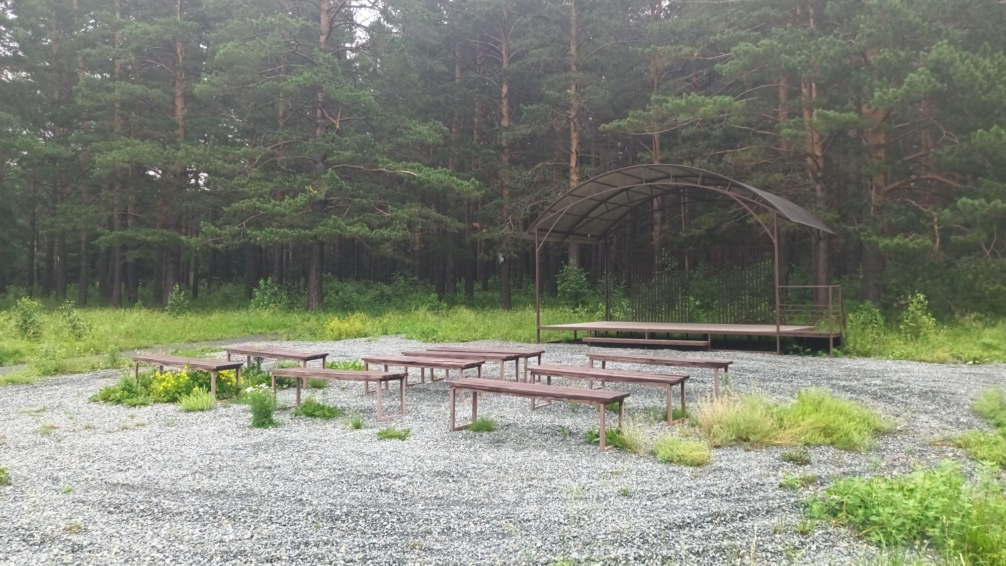 The image depicts an outdoor amphitheater situated in a forest clearing within the city of Abaza, Russia. The scene is dominated by a dense pine forest that forms a backdrop to the amphitheater. The ground is covered with gravel. In the foreground and mid-ground, multiple rows of wooden benches are arranged, facing a raised wooden stage with a covered roof structure. The stage has a railing and steps on its right side. The overall impression is one of a natural and serene setting, possibly used for performances or gatherings.